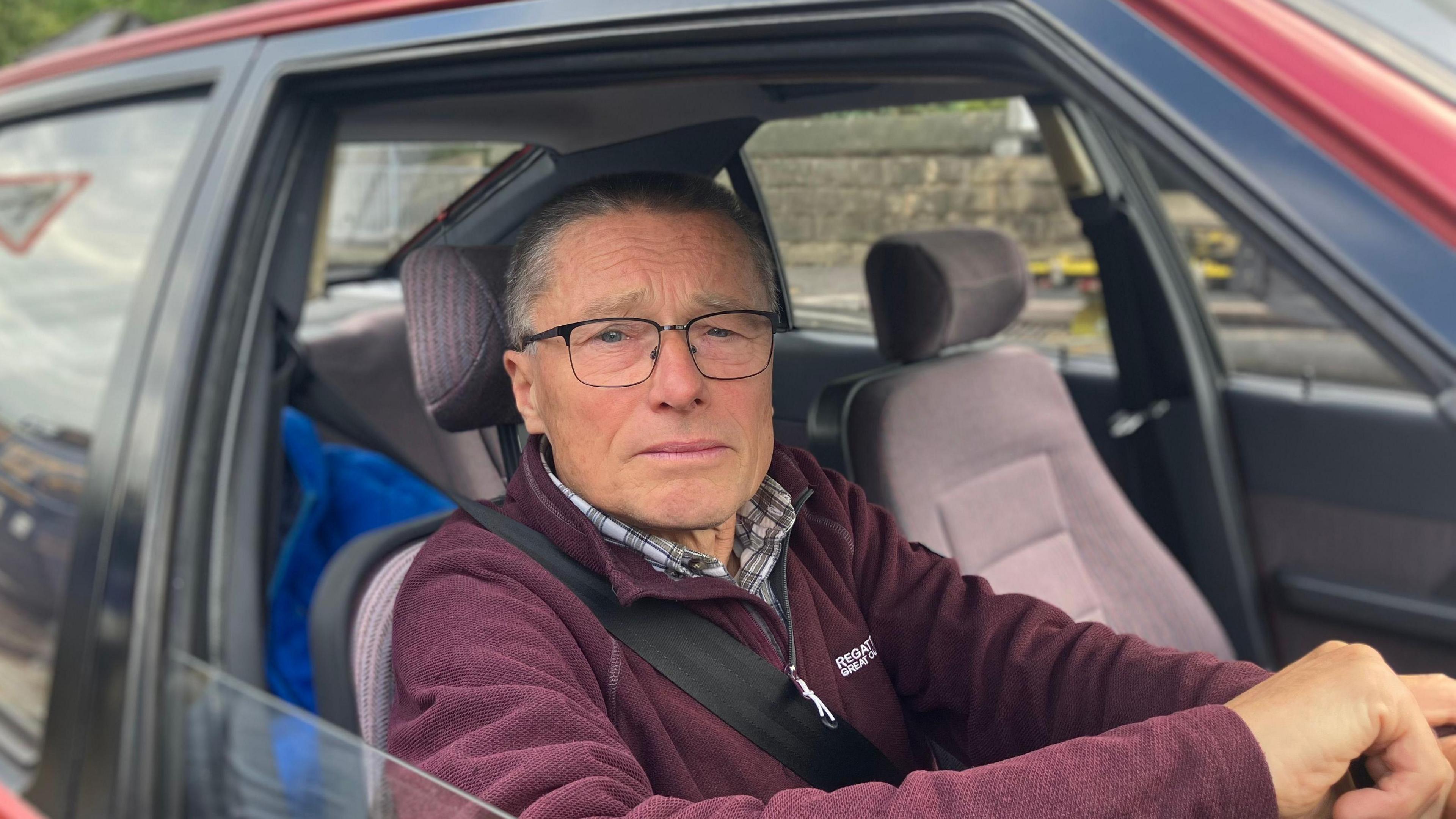 Andrew, a man with short grey hair and glasses sits in the driver's seat of a red car. He is looking into the camera with the car window wound down.