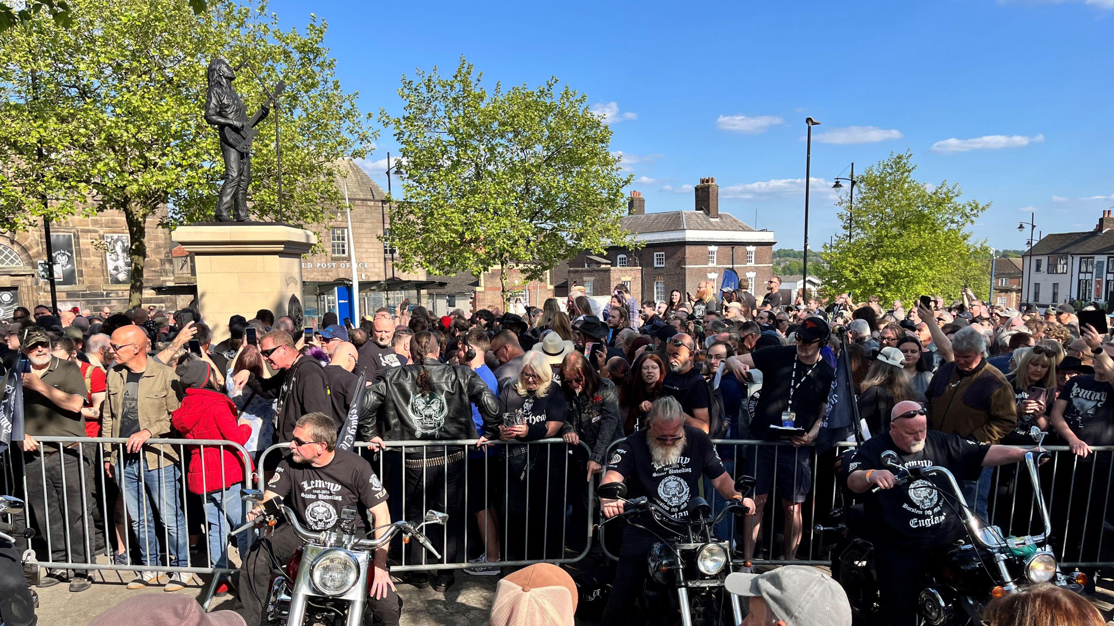 Three men on bikes in front of a railing, behind which is a crowd of people next to the new statue of Lemmy.