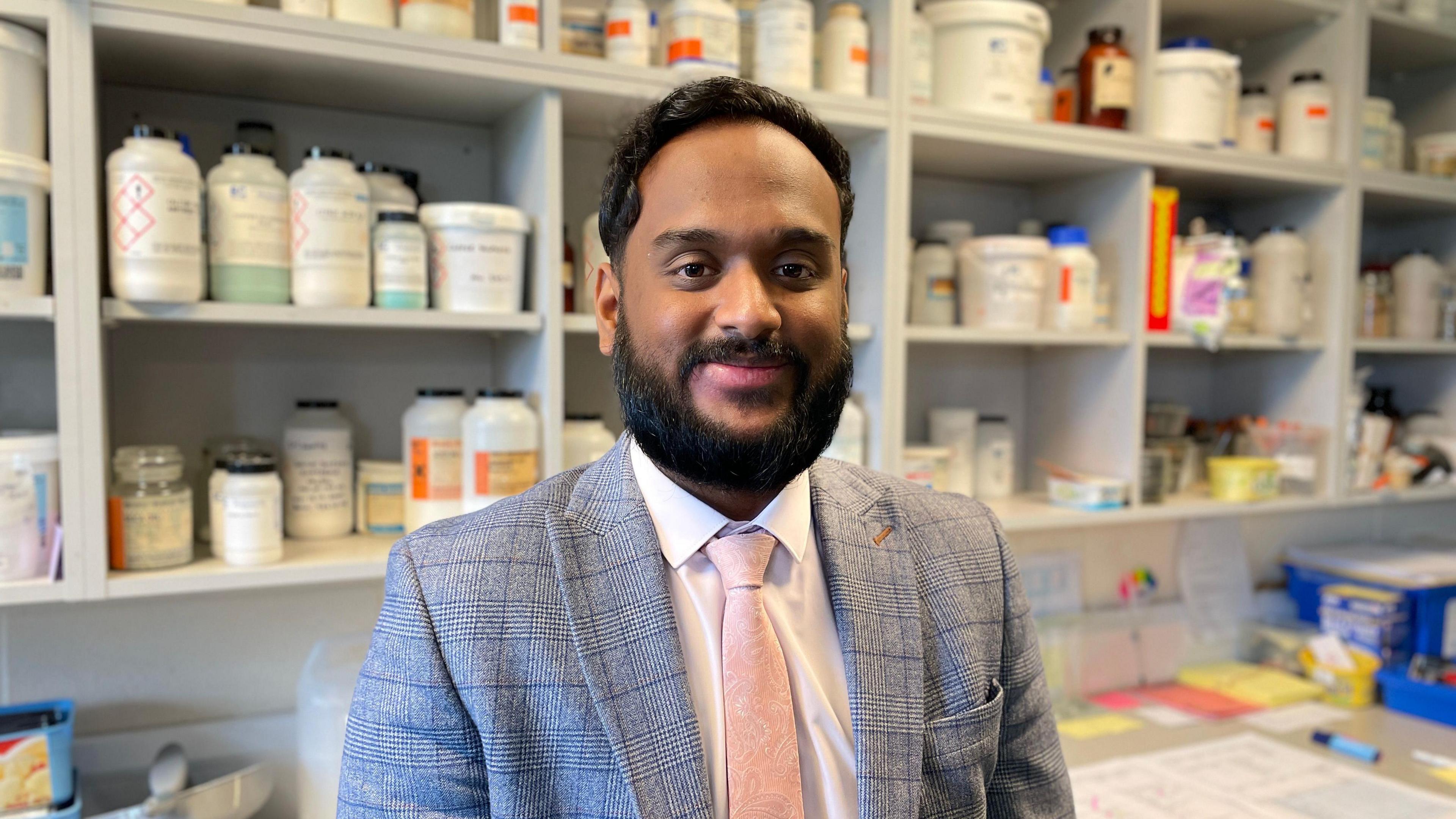 Science teacher Abid Mustafa in the science lab at St Peter’s Catholic School. He is wearing a white shirt, pale pink tie and grey checked suit. Behind him are shelves lined with pots of chemicals. 