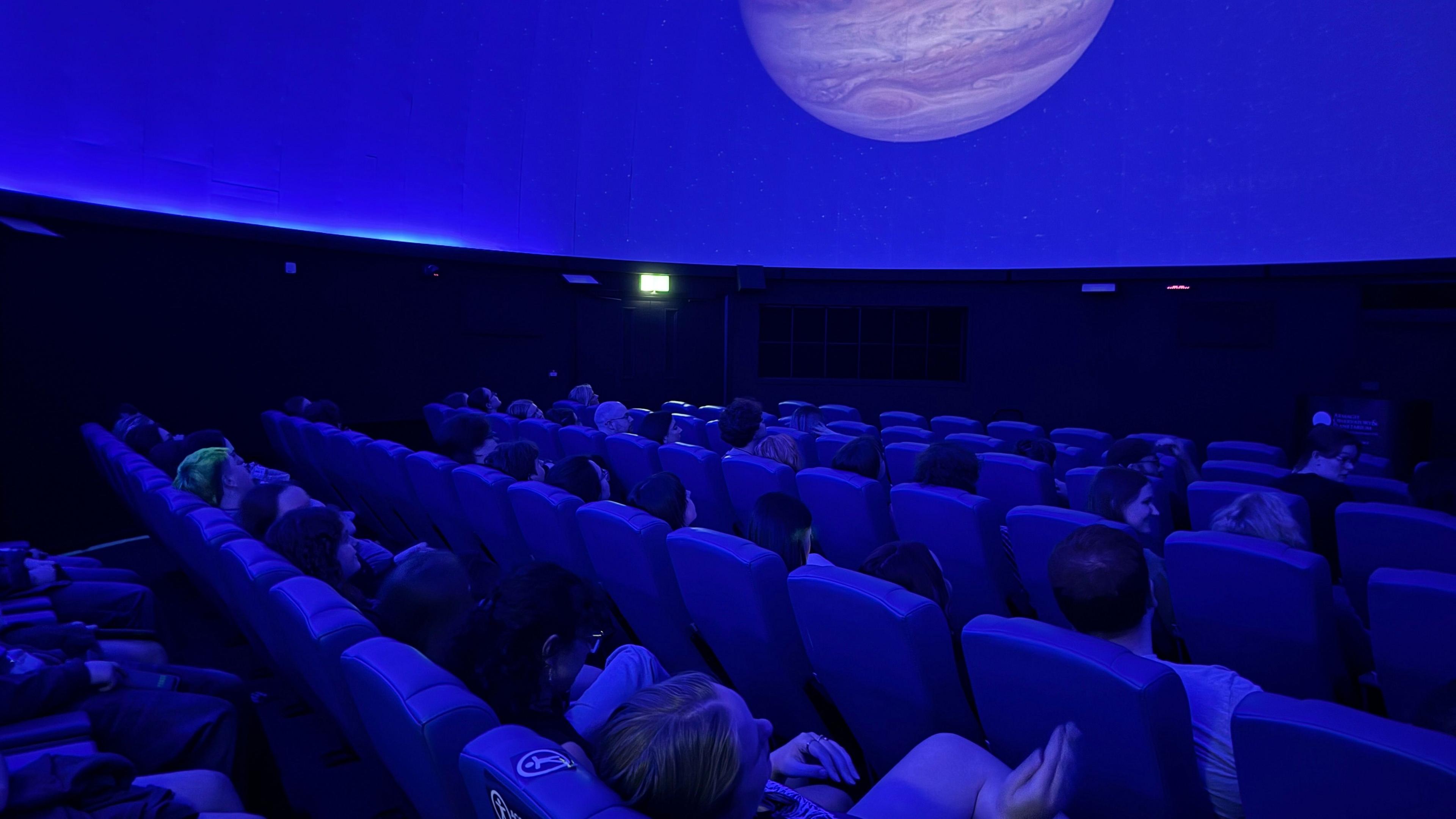 The audience sit in rows of seats in the planetarium's main dome room. The room is lit blue.