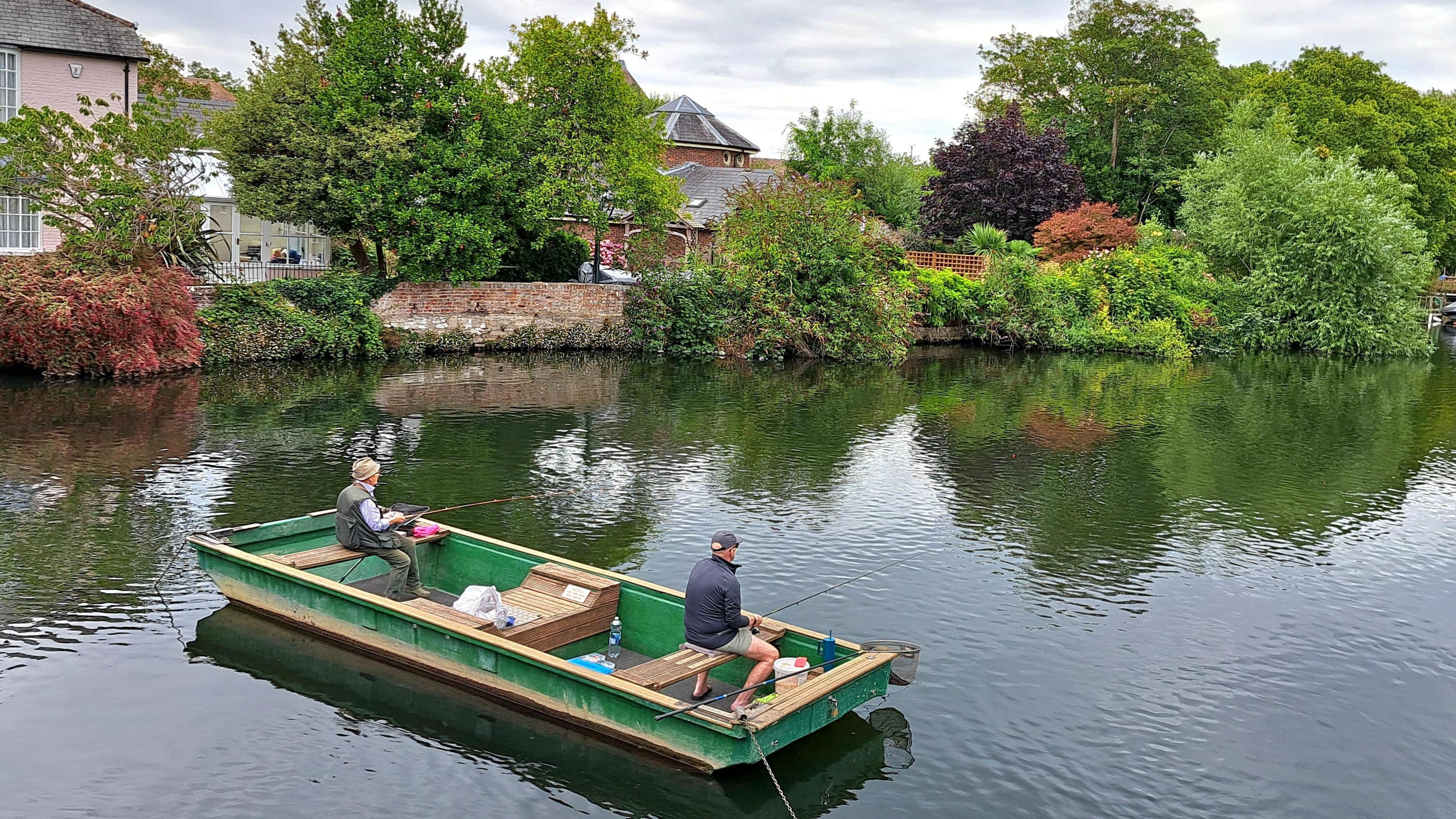 Two fisherman sit in a floating flat boat on the water. Each has a fishing rod and is looking across the surface of the river. Behind them are trees and houses and a red brick wall on the water's edge.