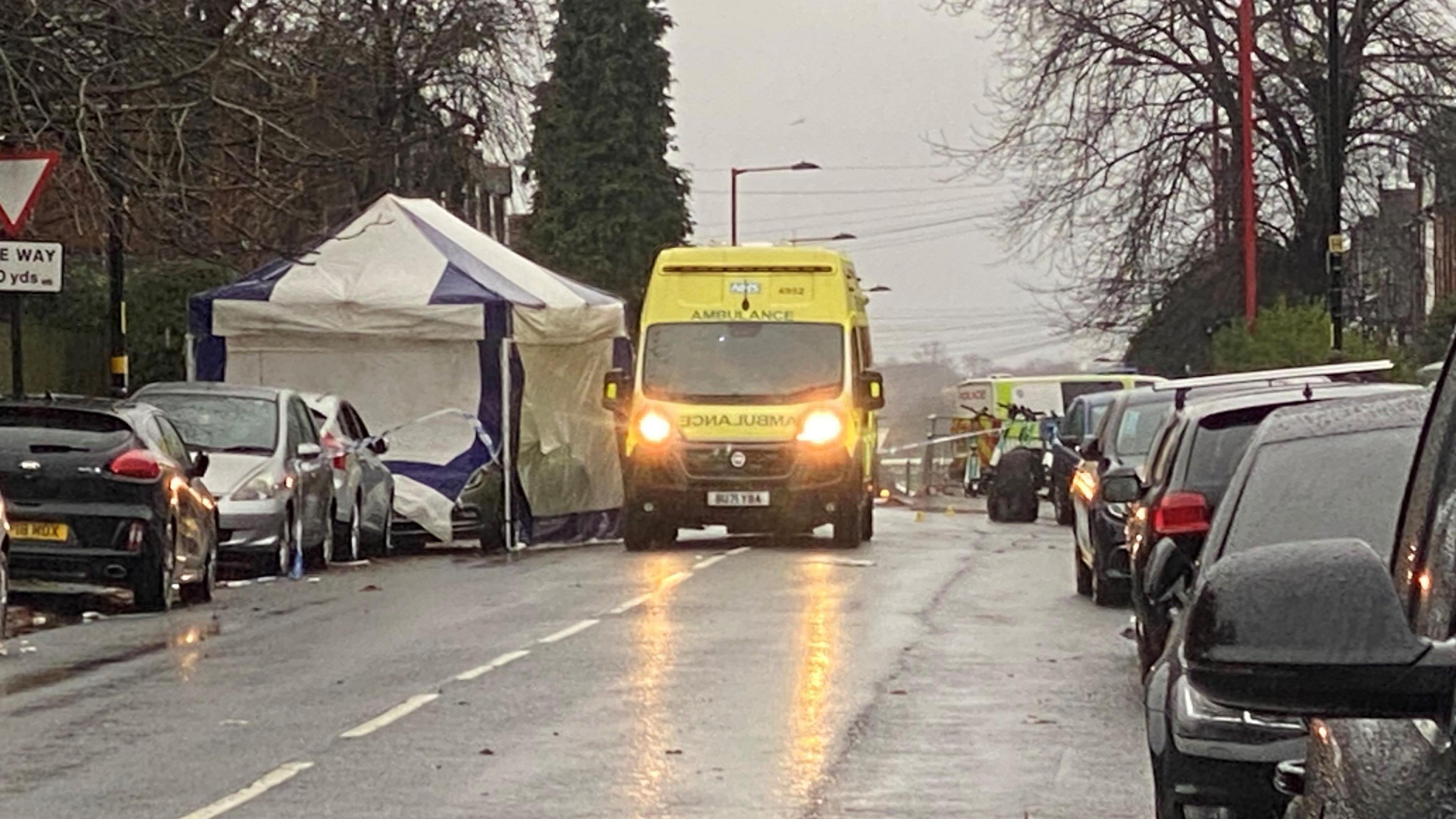 A rain-soaked road, there are park cars either side and a yellow ambulance with its lights on. It is next to a blue and white tent.