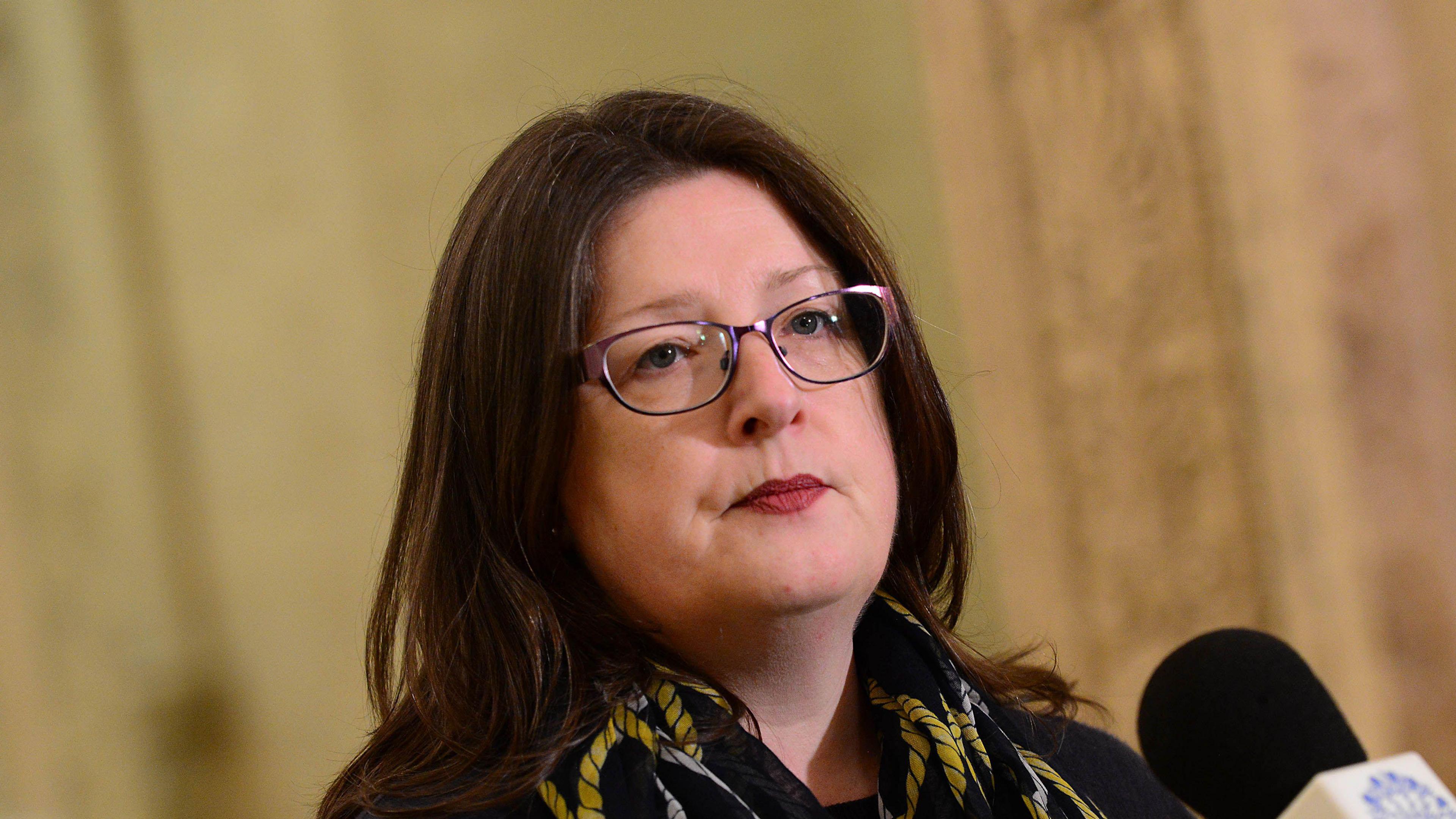 Kellie Armstrong speaking in front of a mic in the Great Hall at Stormont. She has long brown hair, wearing glasses, a back top and silk scarf.