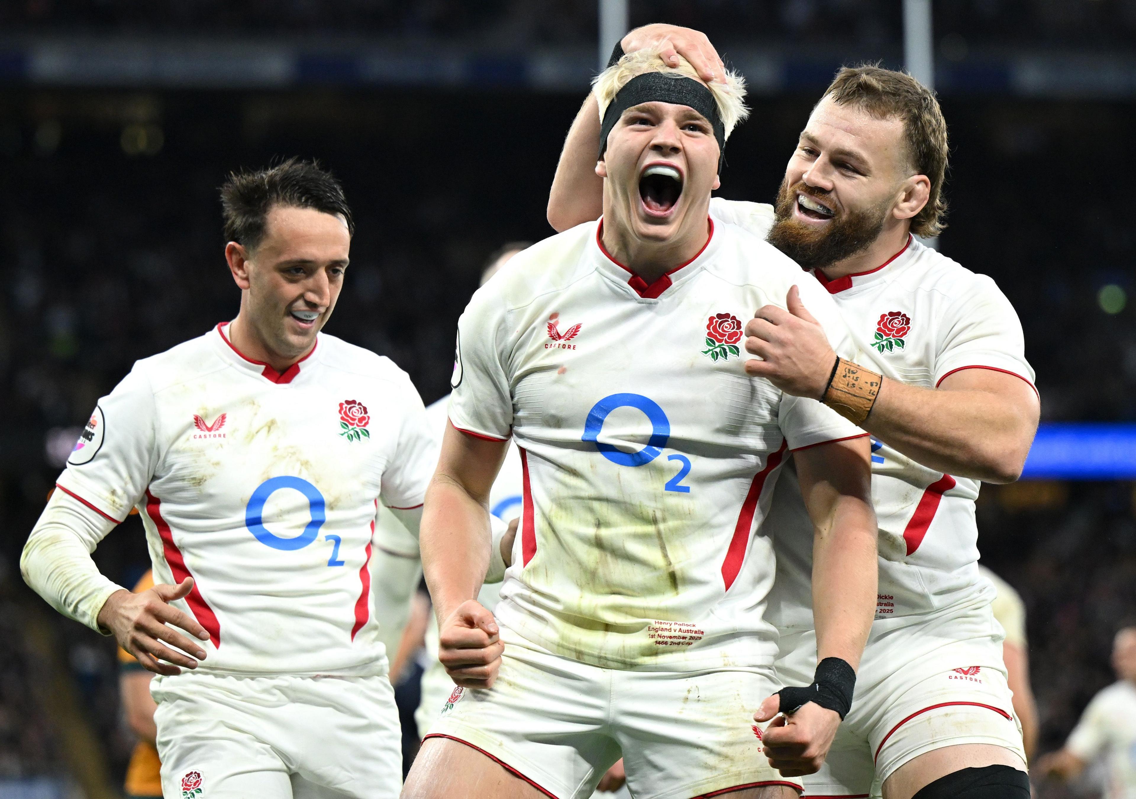 Three rugby players wearing white jerseys with red accents and visible sponsor logos stand on the field during a match. The central player grips the jersey near the waist, while teammates gather close behind. Stadium lights and blurred crowd fill the background.