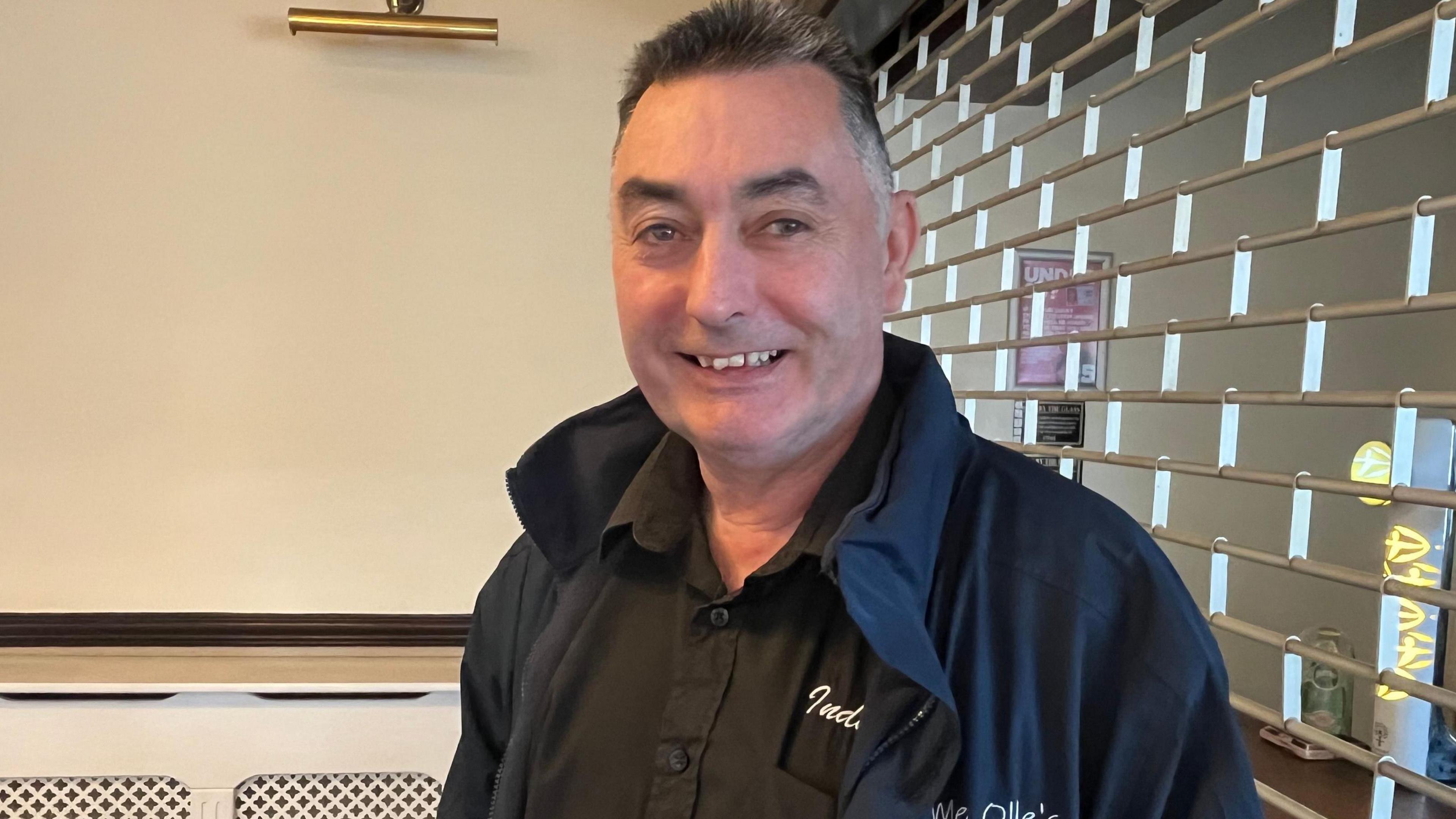 A smiling Danny Brookes dressed in a black shirt and blue jacket looking towards the camera with a metal shutter in the background