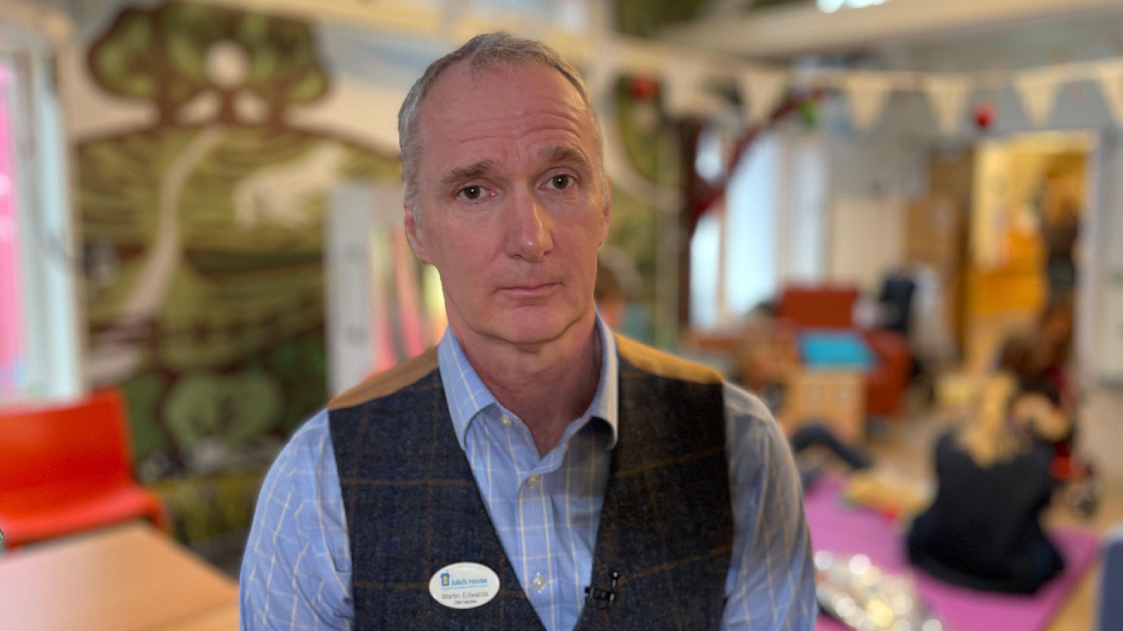 Close up of Martin Edwards' face. He has short grey hair and is wearing a blue shirt while standing inside his hospice. 