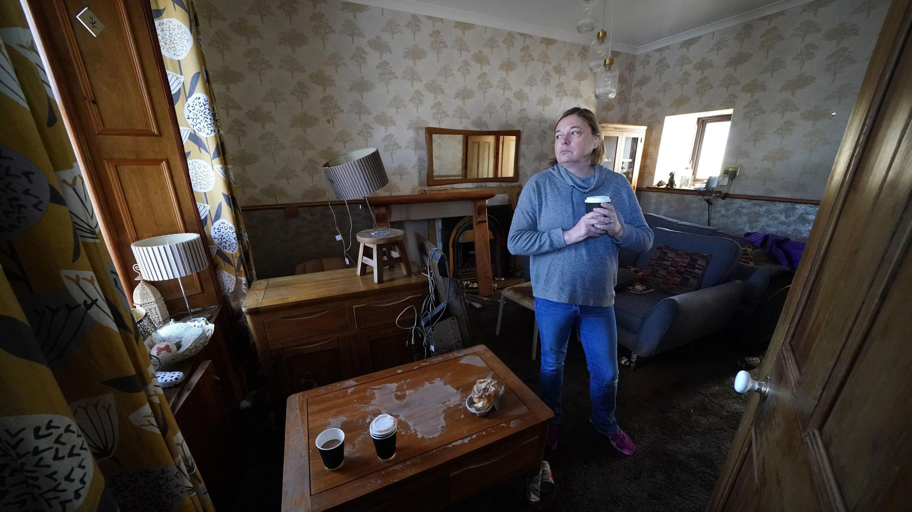 Kim Clark holds a takeaway coffee cup and looks to the side while standing in a flood damaged living room