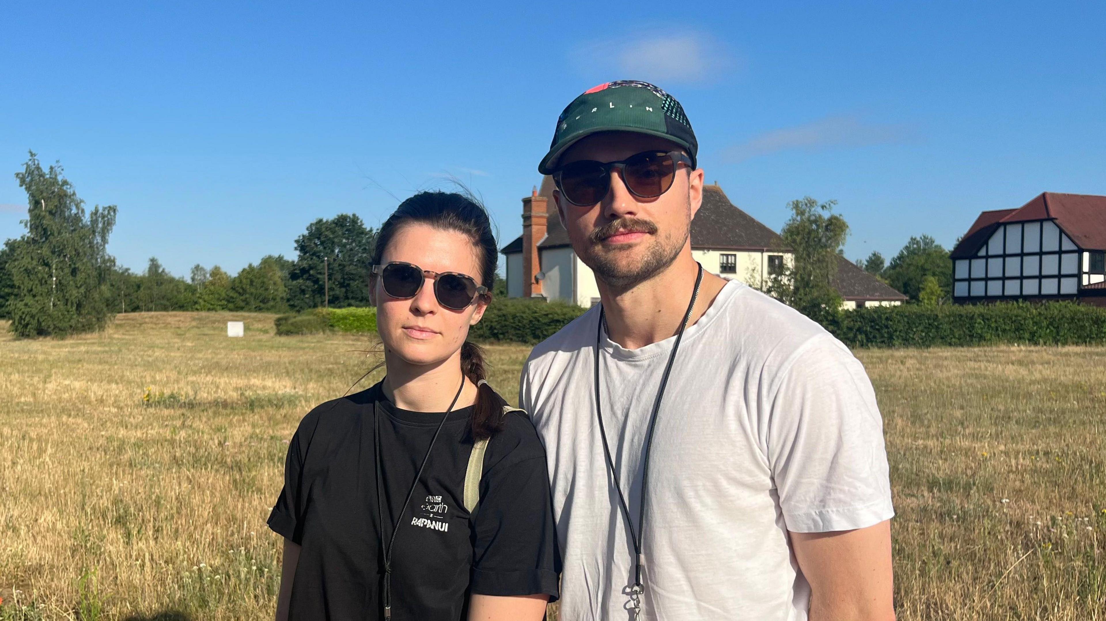 Jess and Tim Roland standing in a field with two detached houses behind them. Jess is wearing dark glasses and a black T-shirt and Tim is wearing a green baseball cap, dark glasses and a white T-shirt.