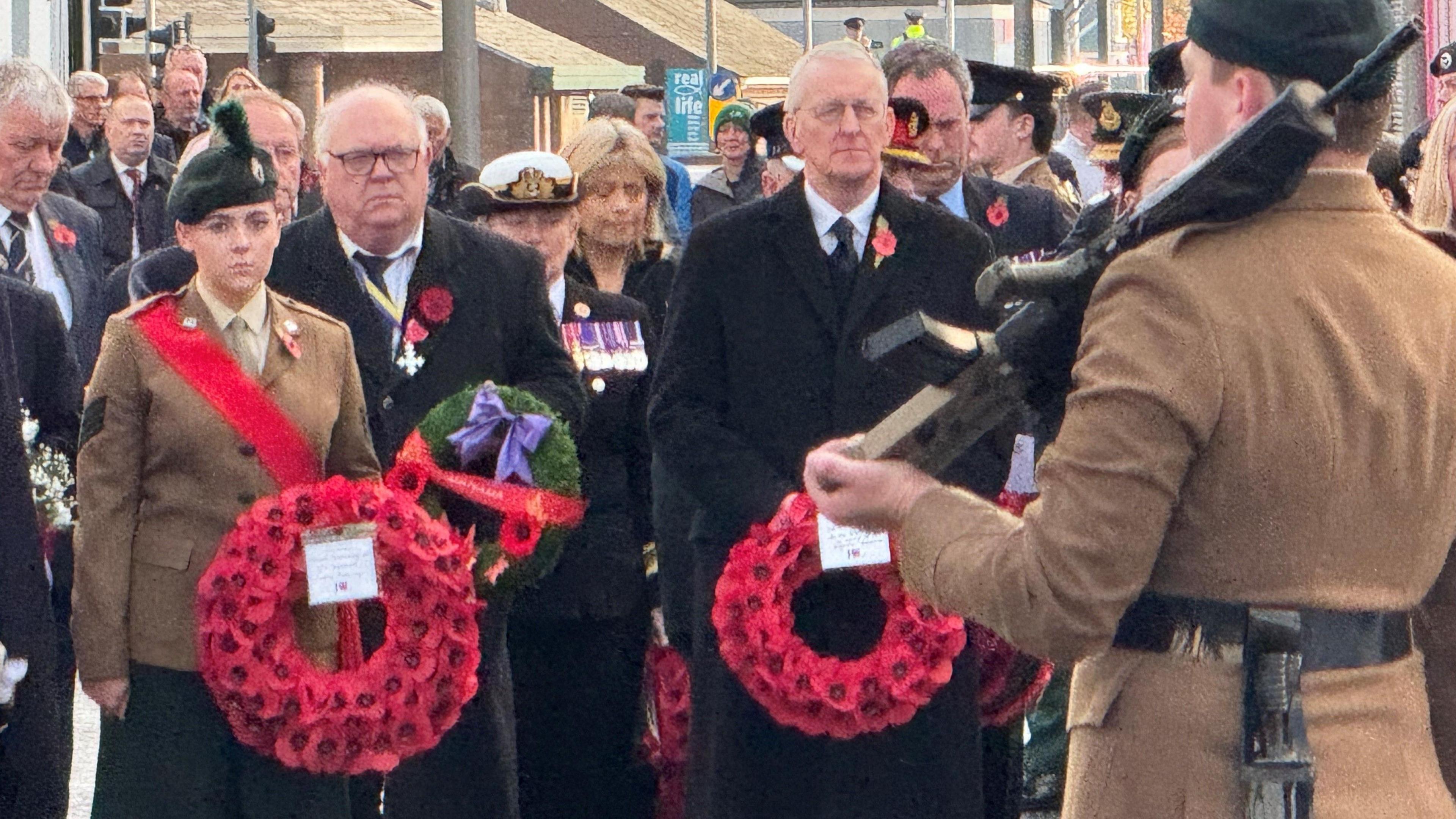 Hilary Benn standing holding a wreath. he is wearing a long black coat with a poppy on it. Beside him a woman in military dress also holding a wrath of poppies. In the foreground a man in military dress holding a rifle facing away from the camera.