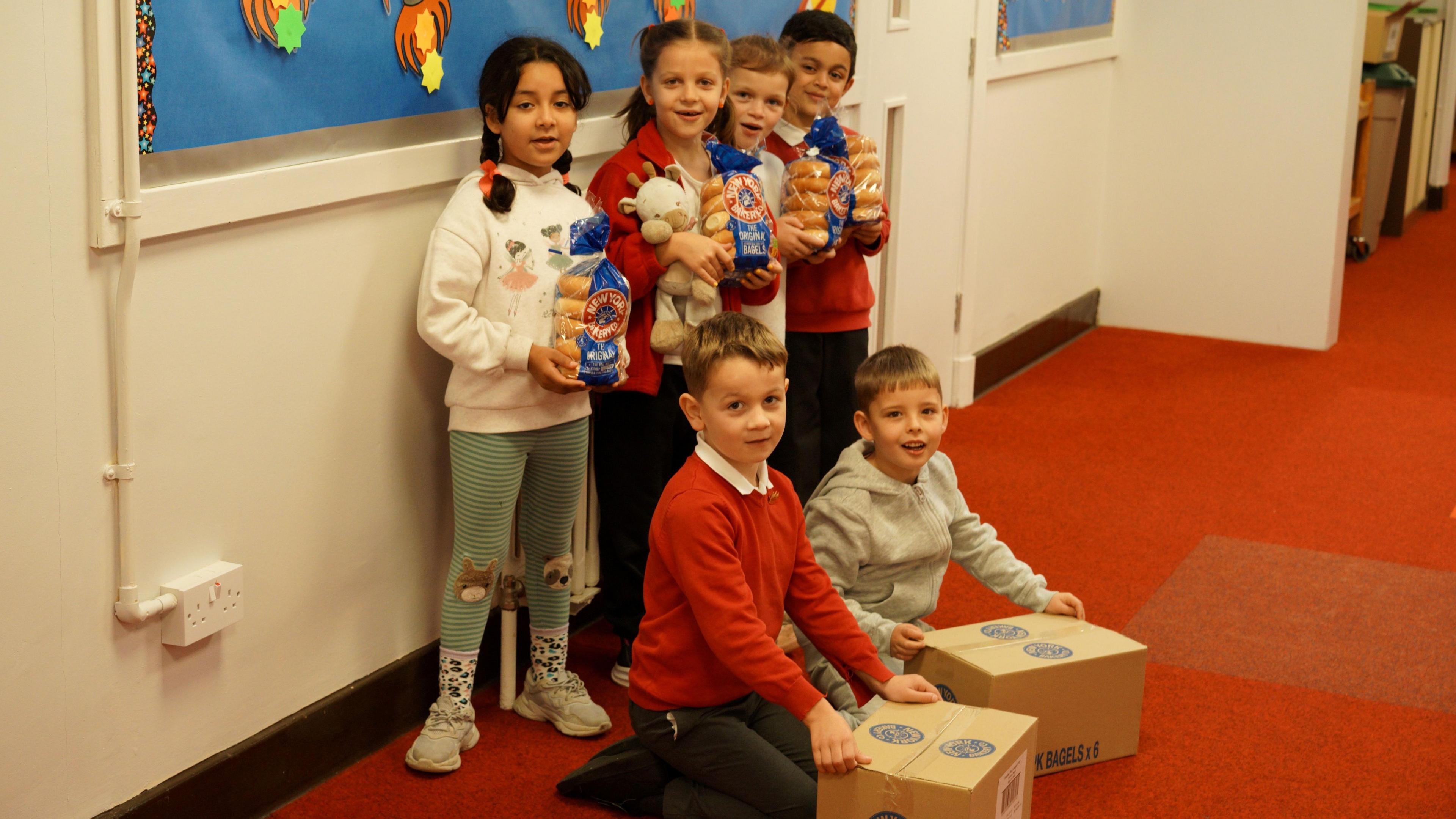 A group of six children, two girls and four boys aged between six and seven, are holding packets or boxes of bagels, they are standing in the school corridor.