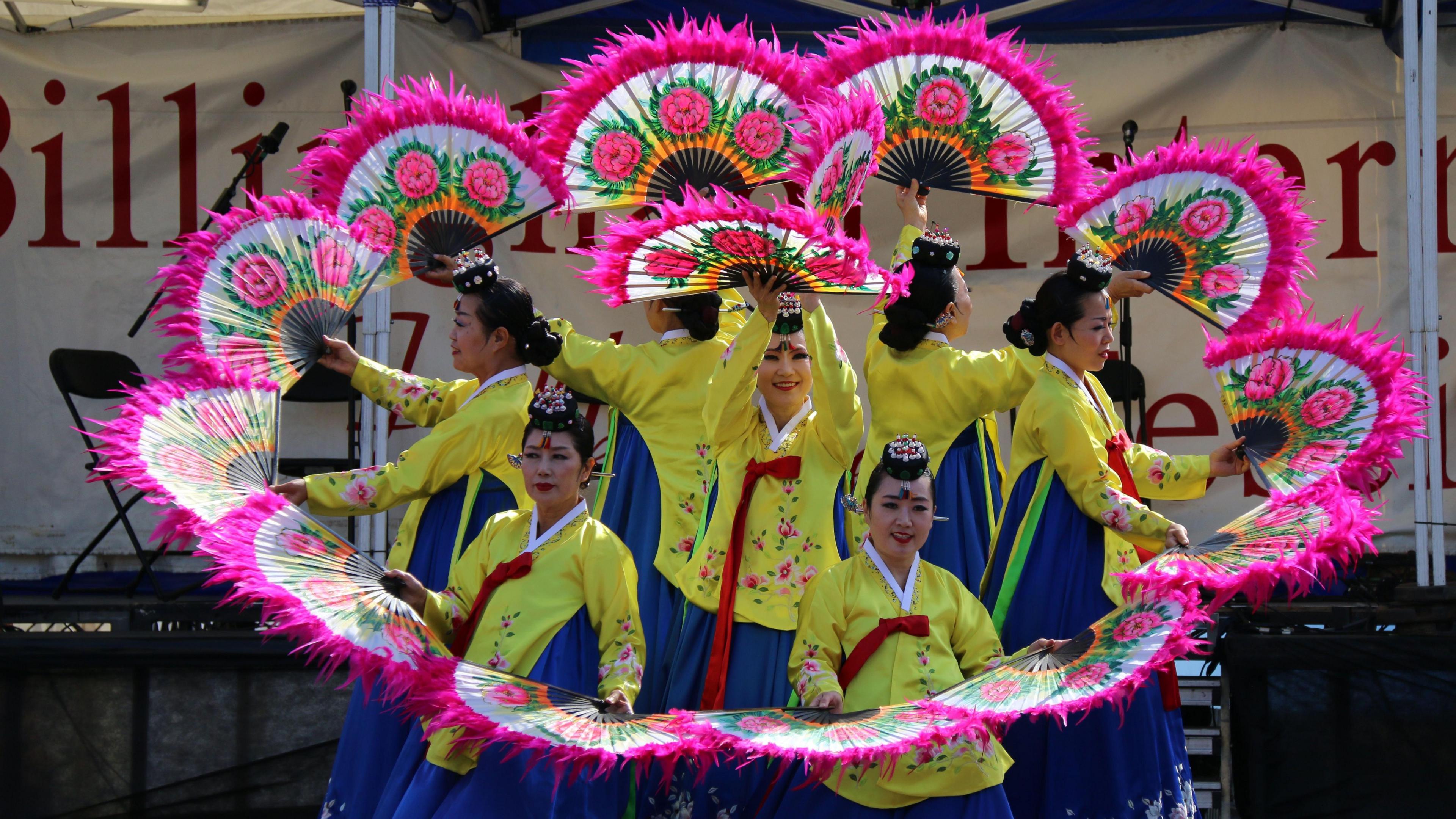 South Korean dancers performing at the festival. They are dressed in yellow and blue dresses and are holding Pink-frilled fans in a circle around themselves.