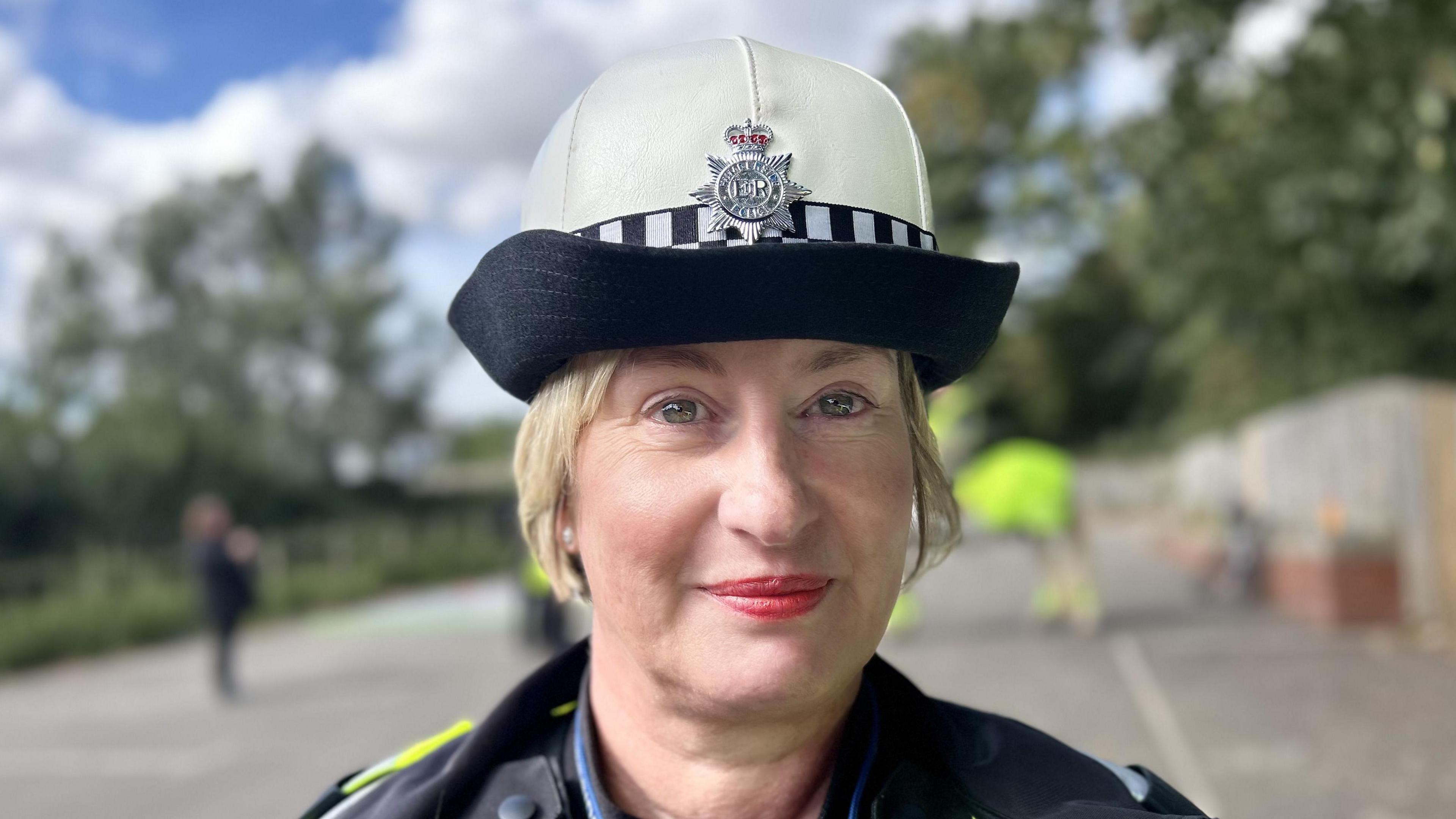 A female police officer with pale skin and blonde hair tied back. She is wearing a police hat with a Humberside Police crest and has red lips and red blusher on. The background of the picture is out of focus, but you can subtly see she's in a car park.