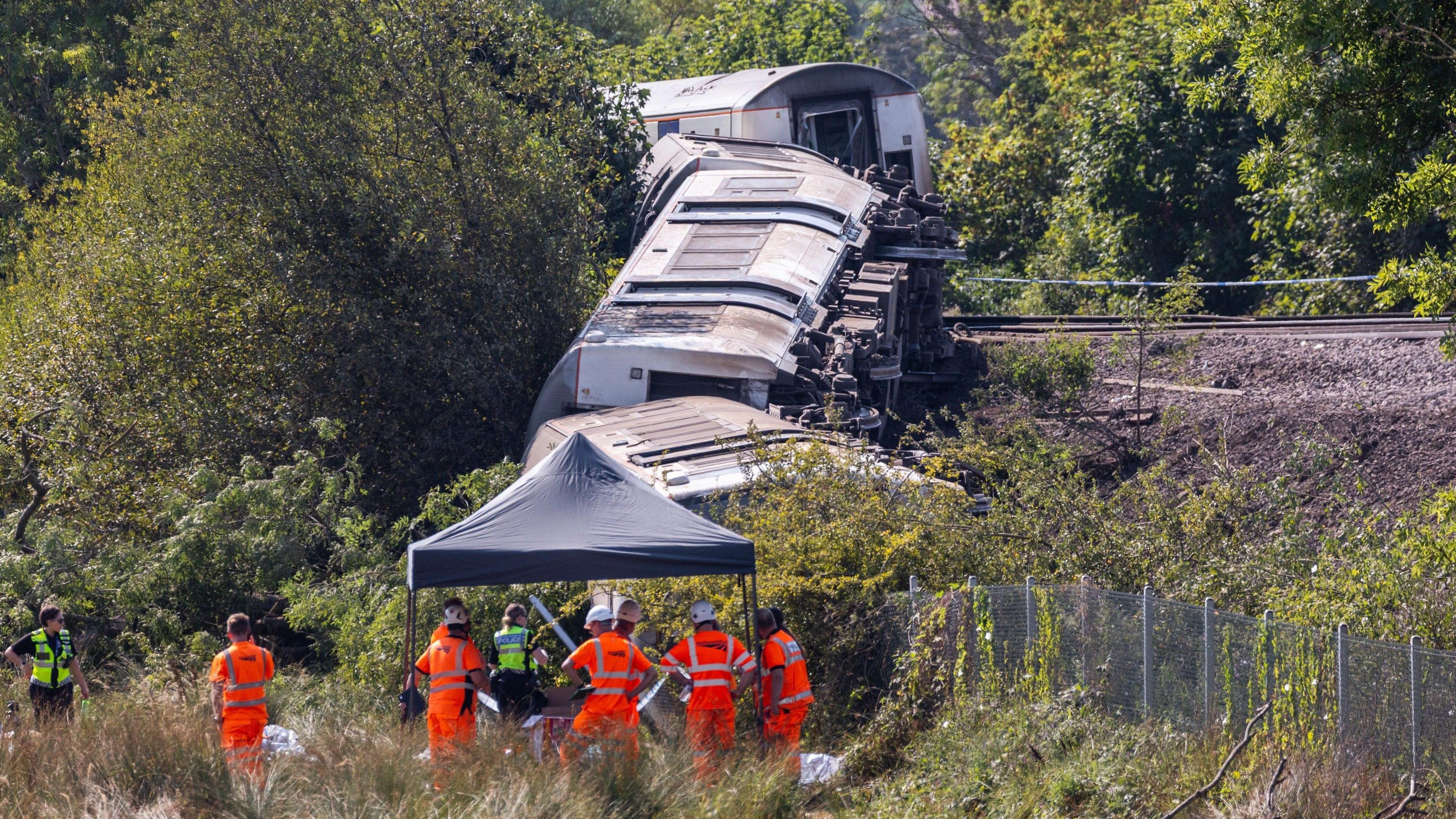 Railway carriages on their side, stretching from a railway line down the side of a small embankment. Emergency workers crowd at the bottom of the embankment.