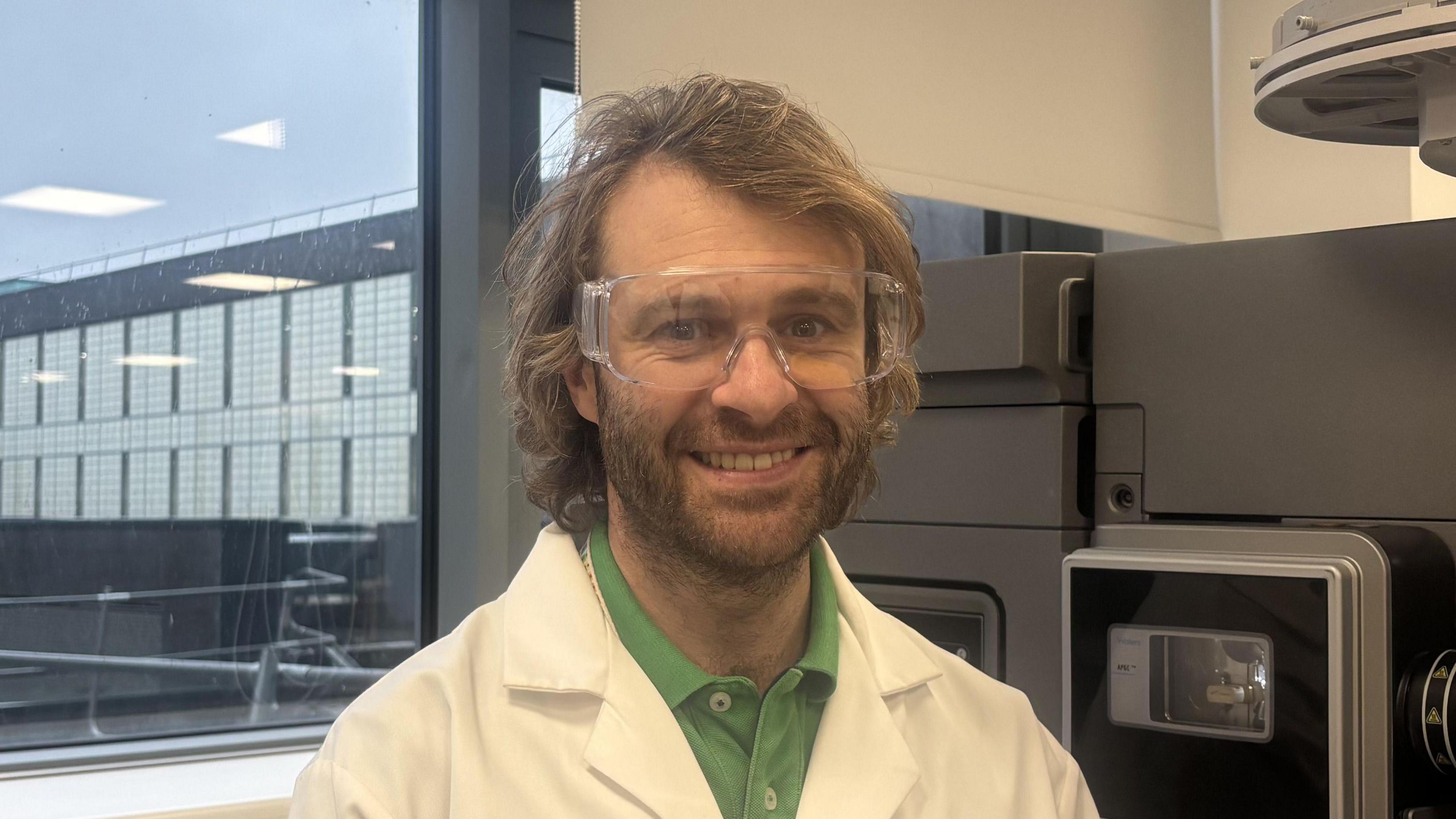 Dr David Megson smiles at the camera. He is in a lab and there is a metal machine behind him. He has long shoulder-length curly brown hair and facial hair. He is wearing a green polo top and a white lab coat and protective glasses. 