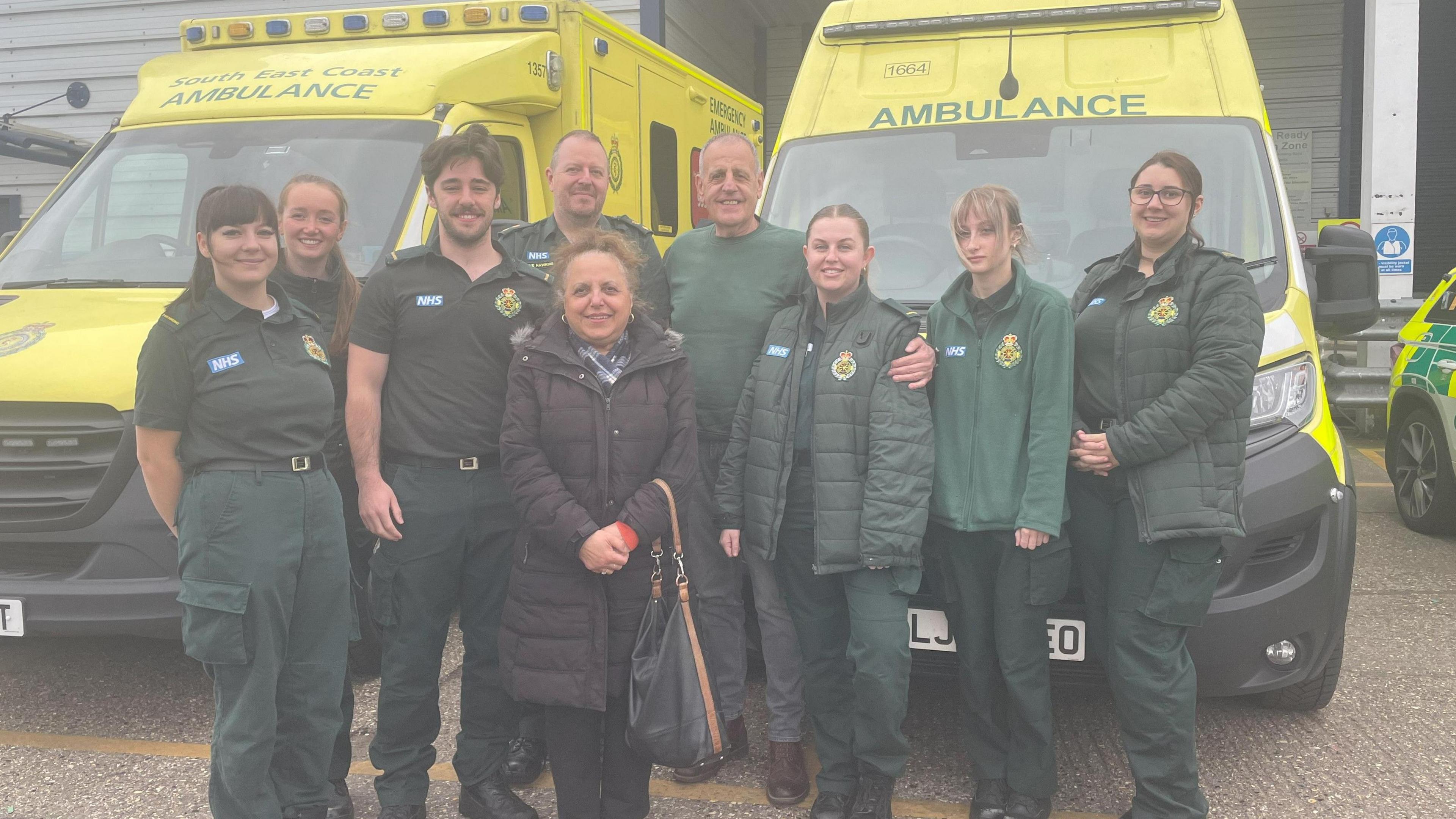 Staff at South East Coast Ambulance Service pose for a photograph with Antony Linward. The group are stood in front of two bright yellow ambulances, with the medical team wearing dark green NHS uniforms.