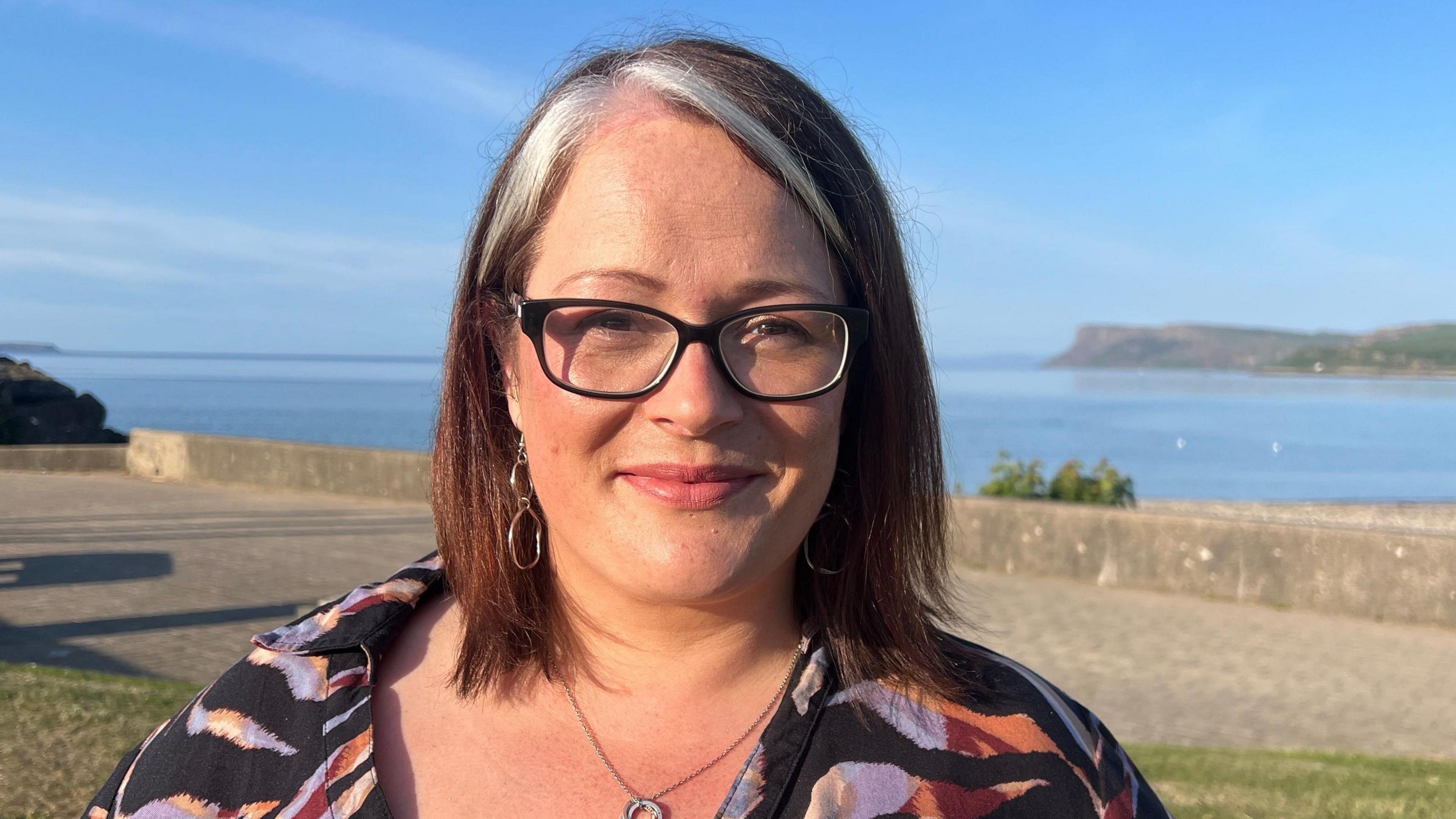 A woman with red hair, with a white streak, and glasses is speaking into a microphone. The background is the sea. She is wearing a floral top.