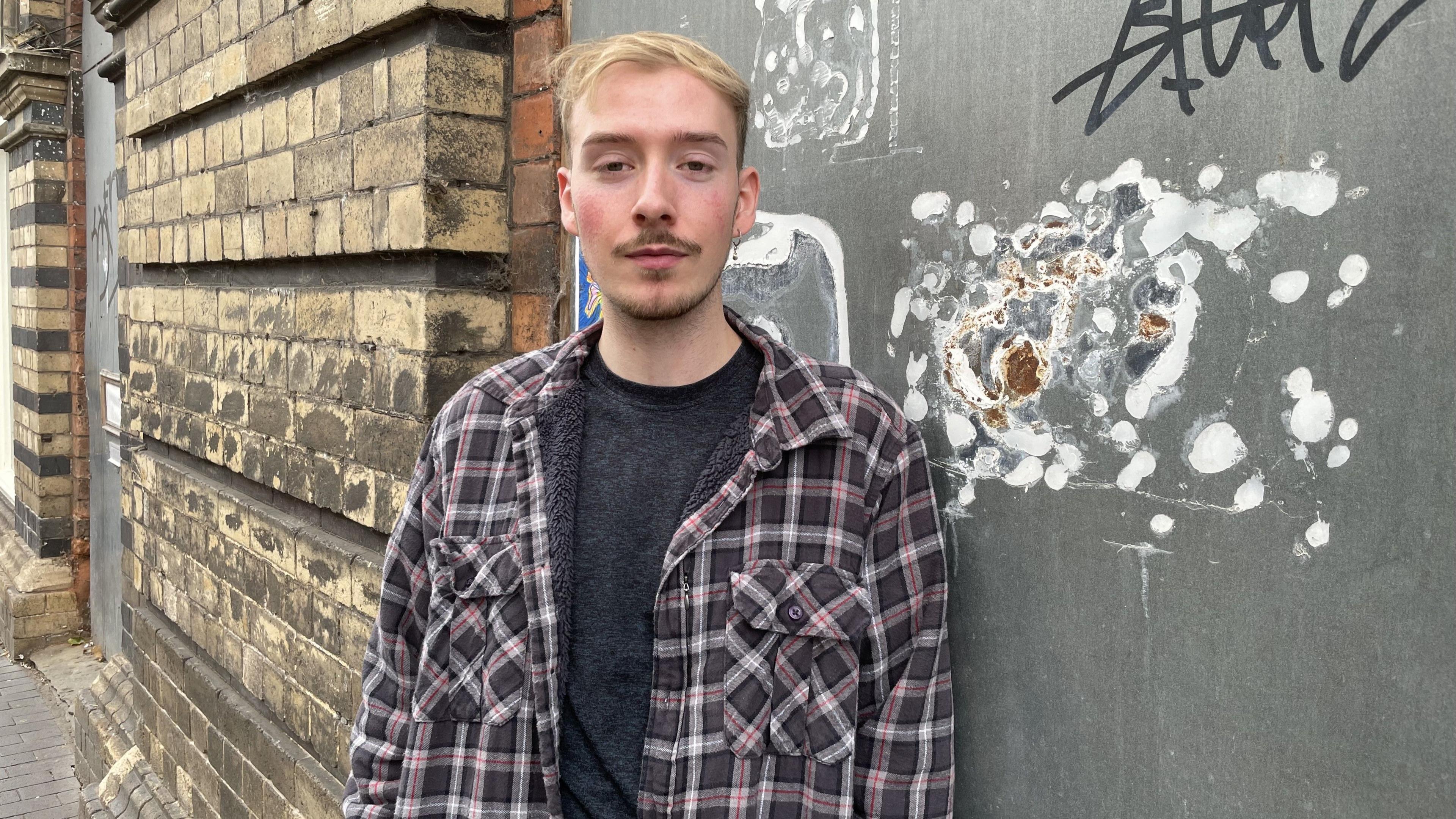 Huw has short blonde hair and brown stubble with a brown moustache. He is wearing a purple, white and grey plaid shirt with a grey top underneath. He is standing in front of a portion of wall that has graffiti on it.