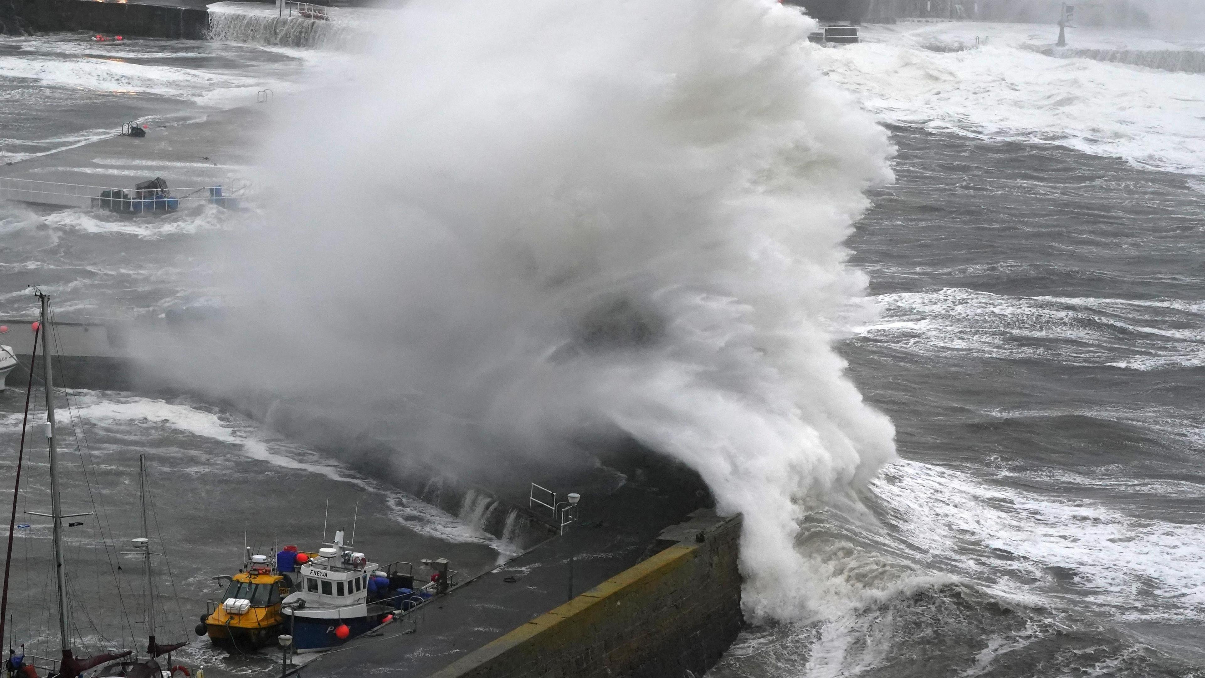 A massive wave blows over the harbour wall at Stonehaven. The plume of water is white, above a grey sea. Two small boats are in the harbour in the foreground.