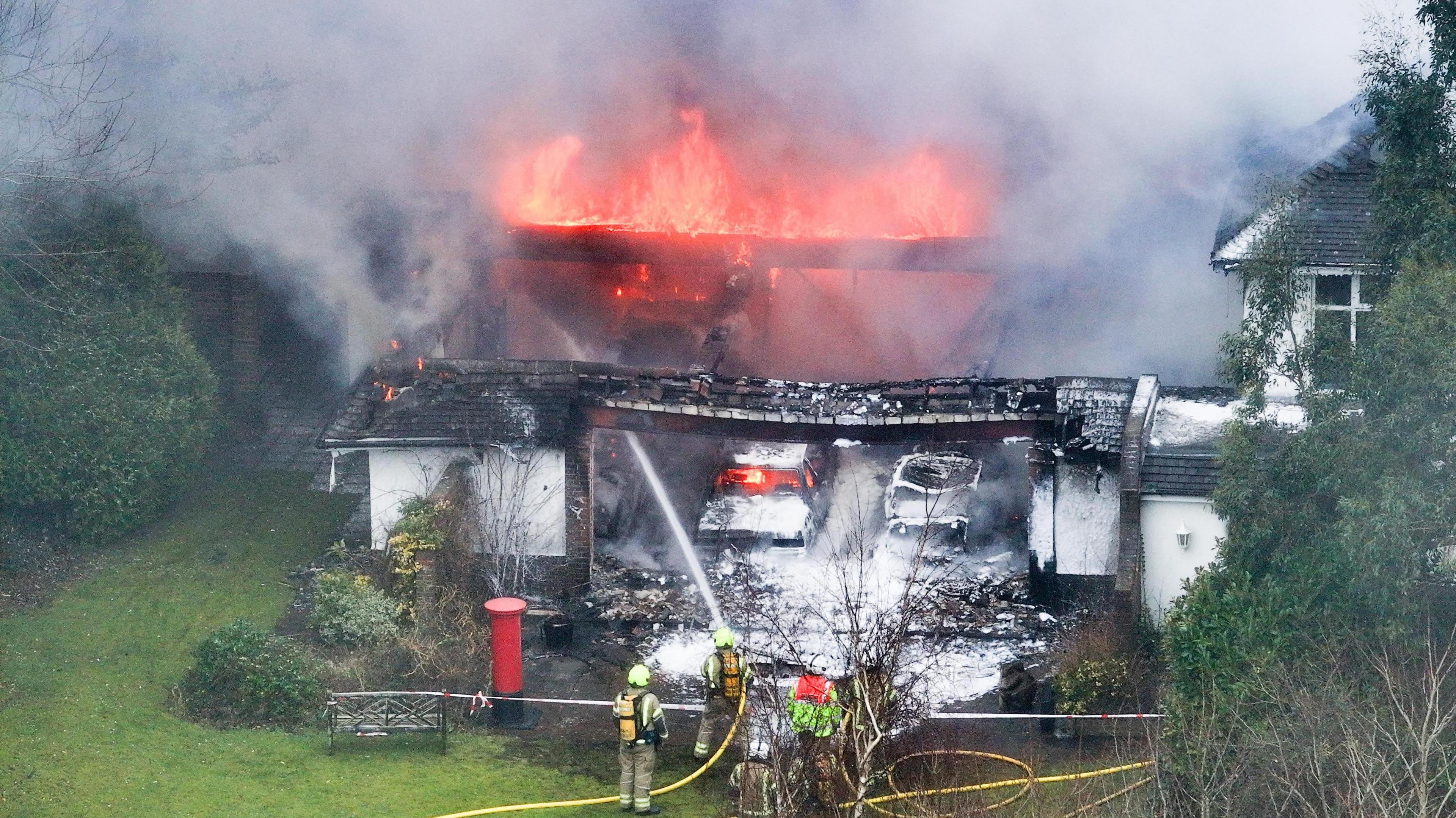 The remains of a one-storey building which has been destroyed by fire. The top of the building is well alight and four fire fighters are on the ground spraying a hose at it