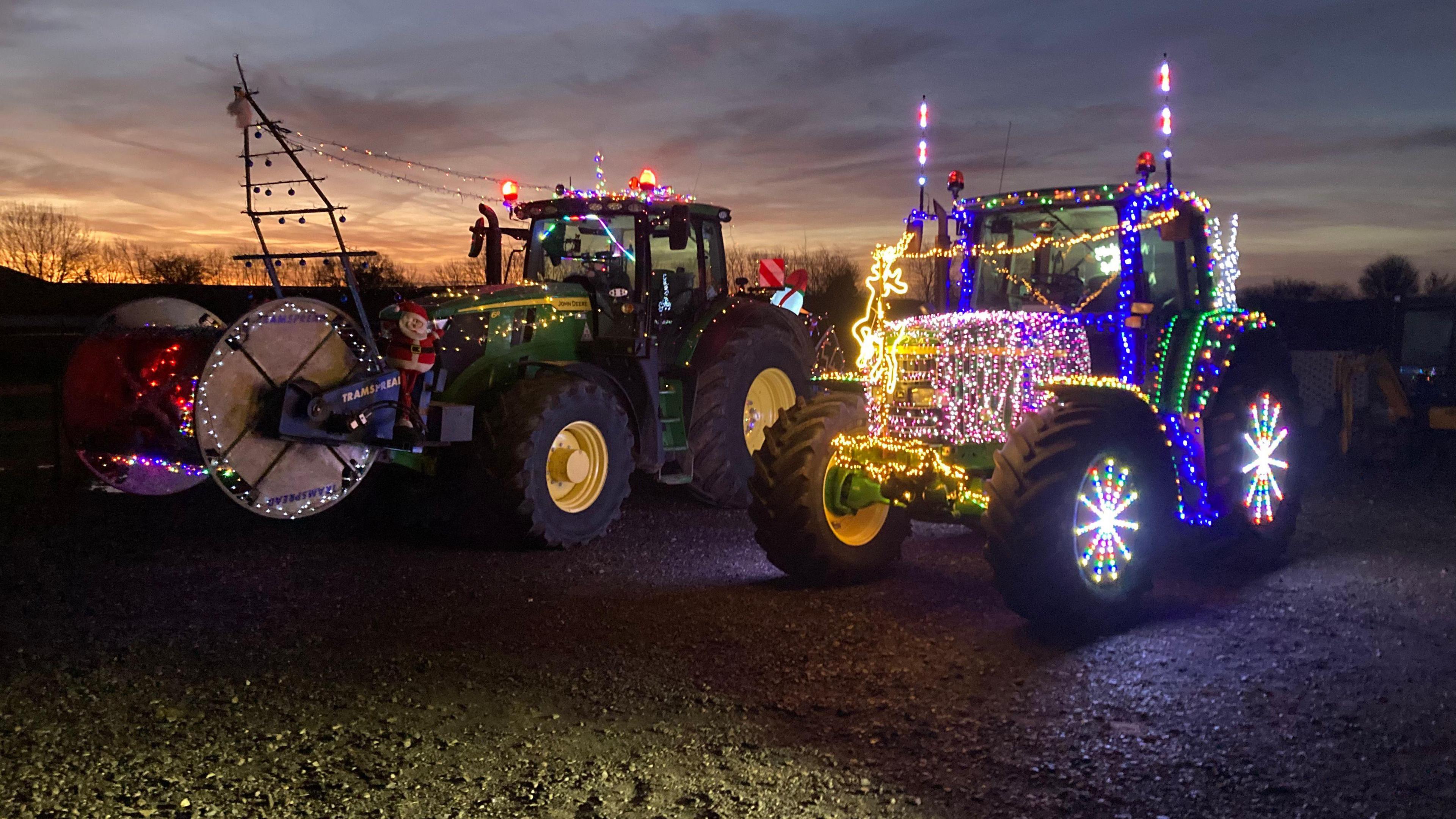 Two tractors in a field decorated with Christmas lights. The sun is rising in the background behind some trees.