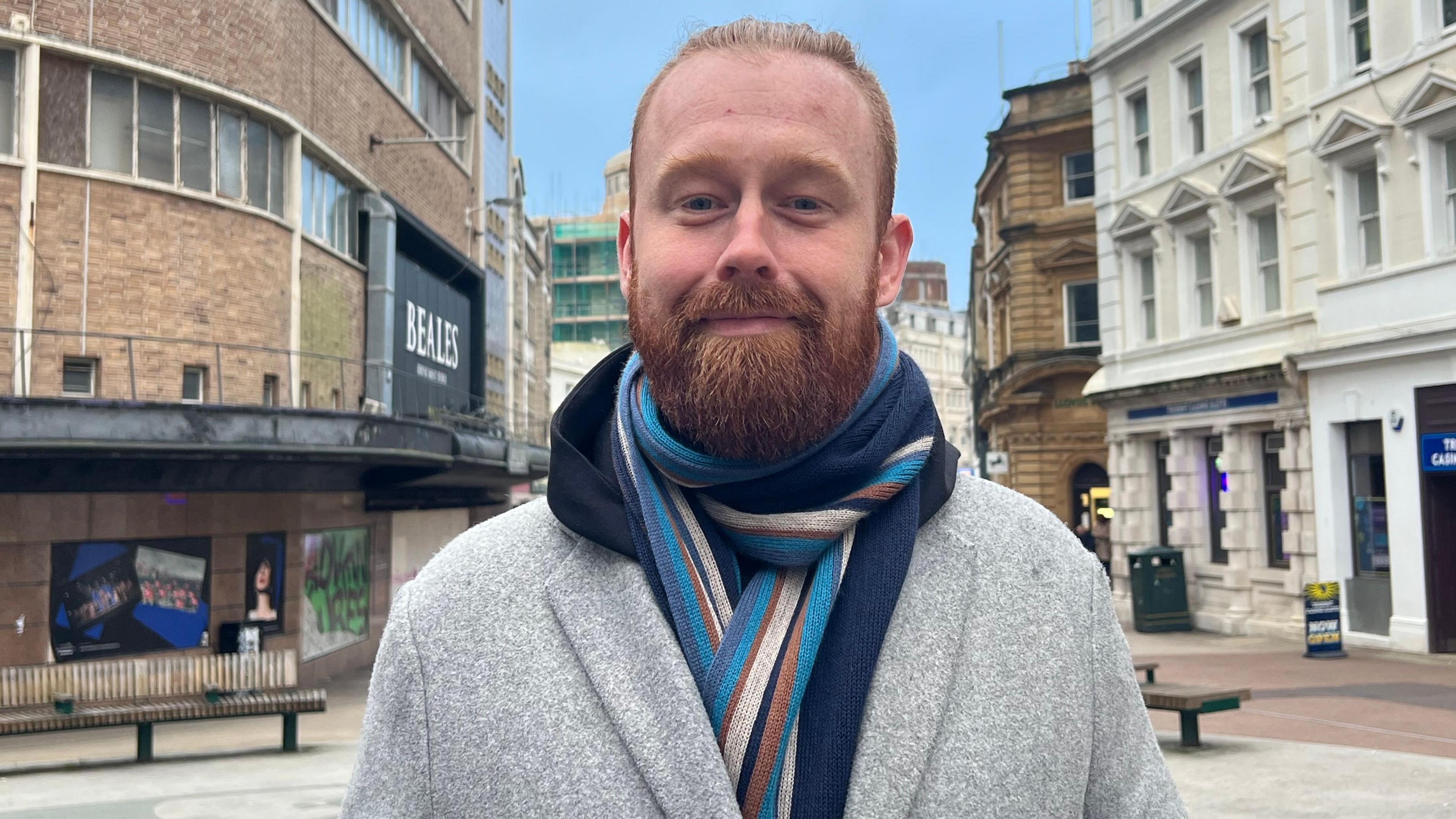 A man stood on a high street in between two buildings. He is wearing a grey coat and a scarf.