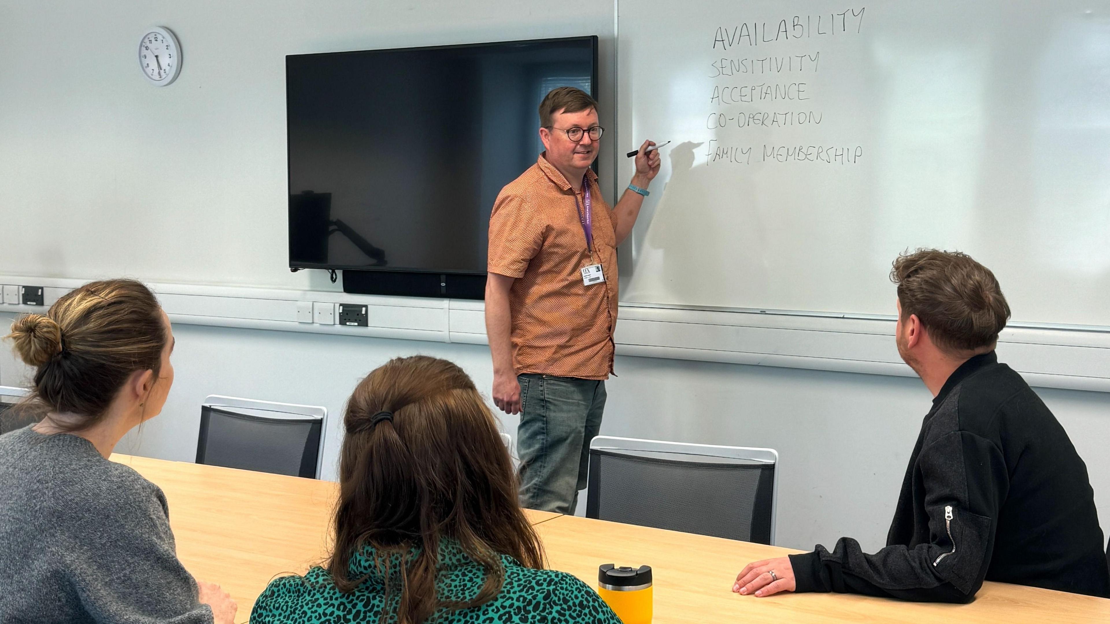 Dr Mark Gregory, wears glasses, a brown shirt and jeans, and stands at a white board with a pen in a classroom. Two women and a man sit with their backs to the camera, looking at Dr Gregory.