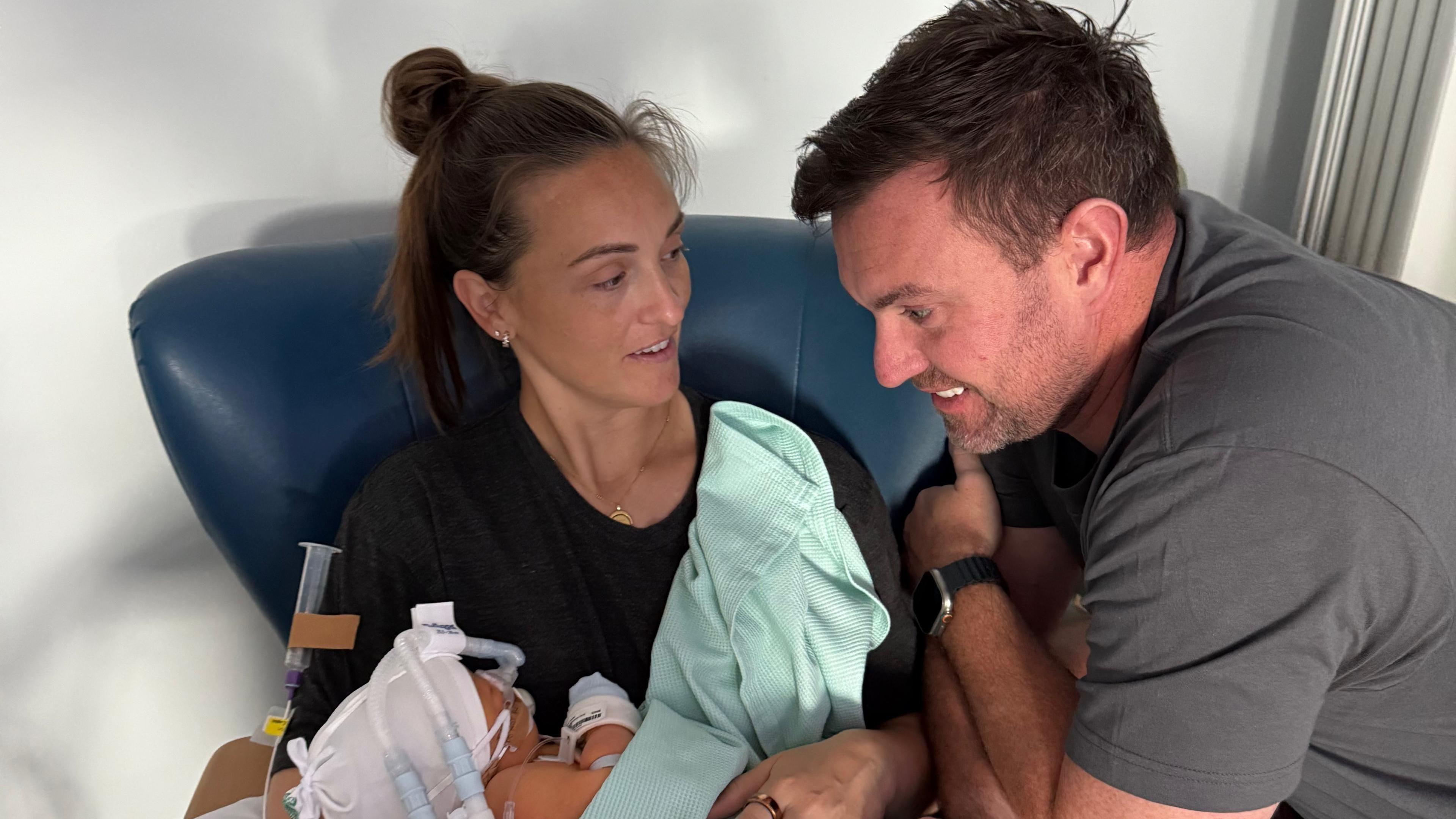 Jade is seated in a hospital chair holding a baby wrapped in a light green blanket. The baby has medical tubes and monitoring devices attached. Chris is leaning close, resting an arm on the chair, with hands touching the seated person’s arm.