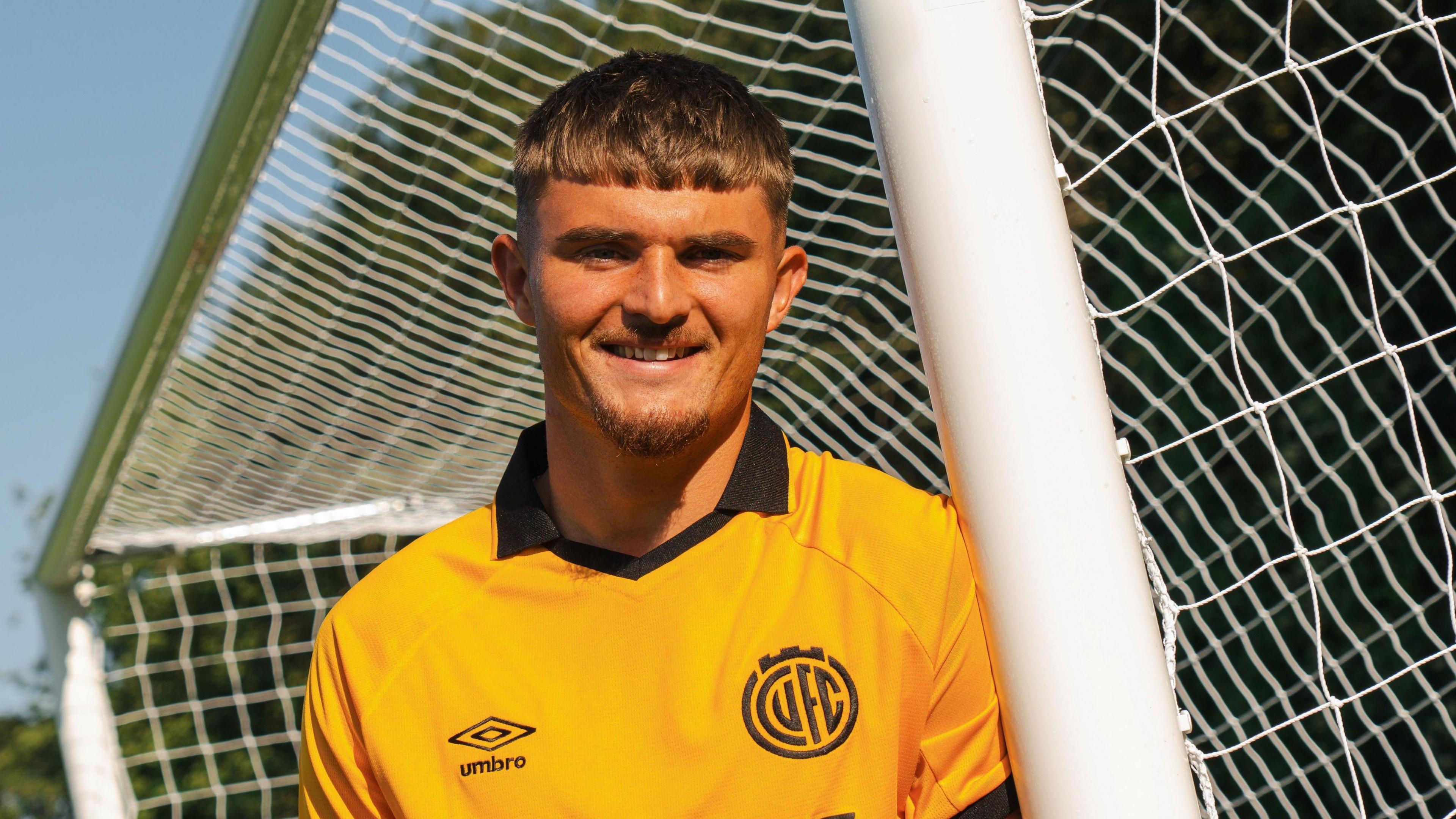 Callum Perry poses against a football goalpost in an amber Cambridge United home jersey.