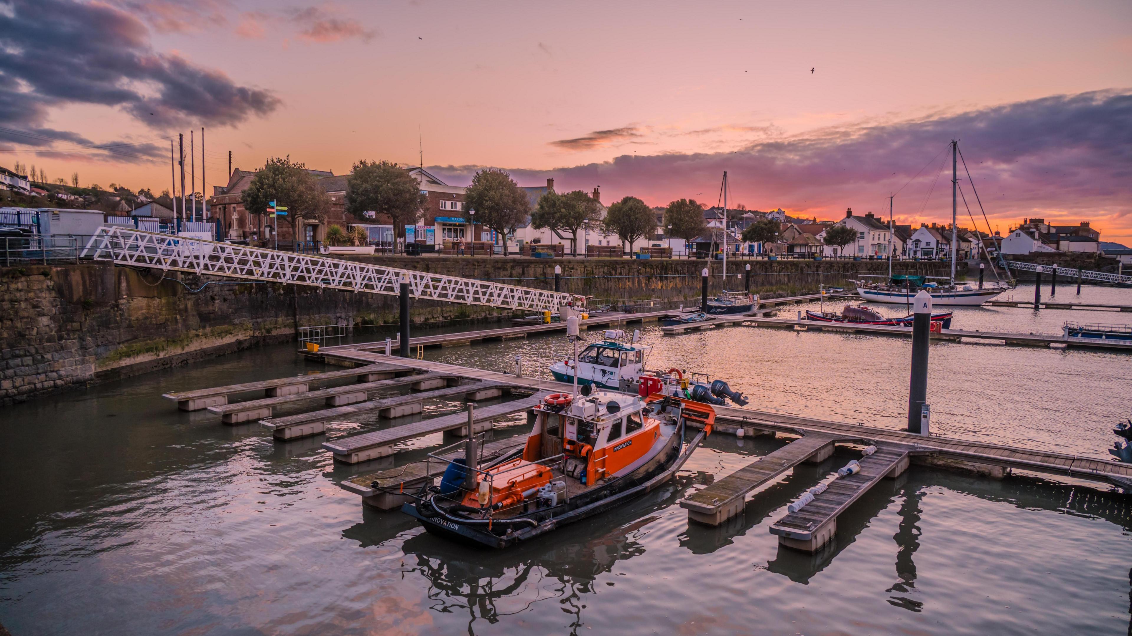 A sunrise is captured over Watchet harbour. There are a few boats by the docks and the sky is pink.