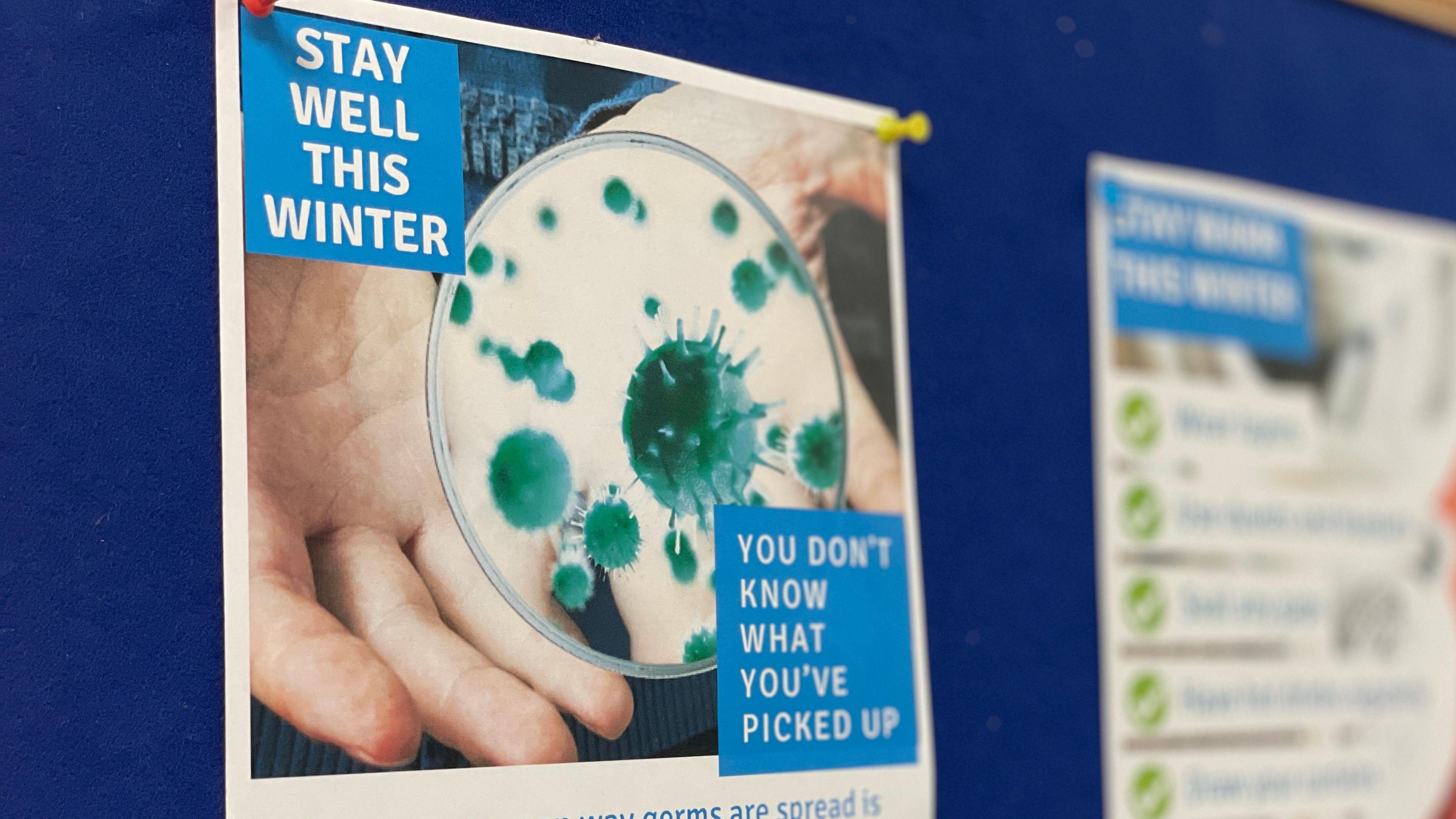 A poster that reads 'stay well this winter' pinned to a blue pinboard. The poster has a photo of a hand and a microscope which is zoomed in on green particles representing germs.