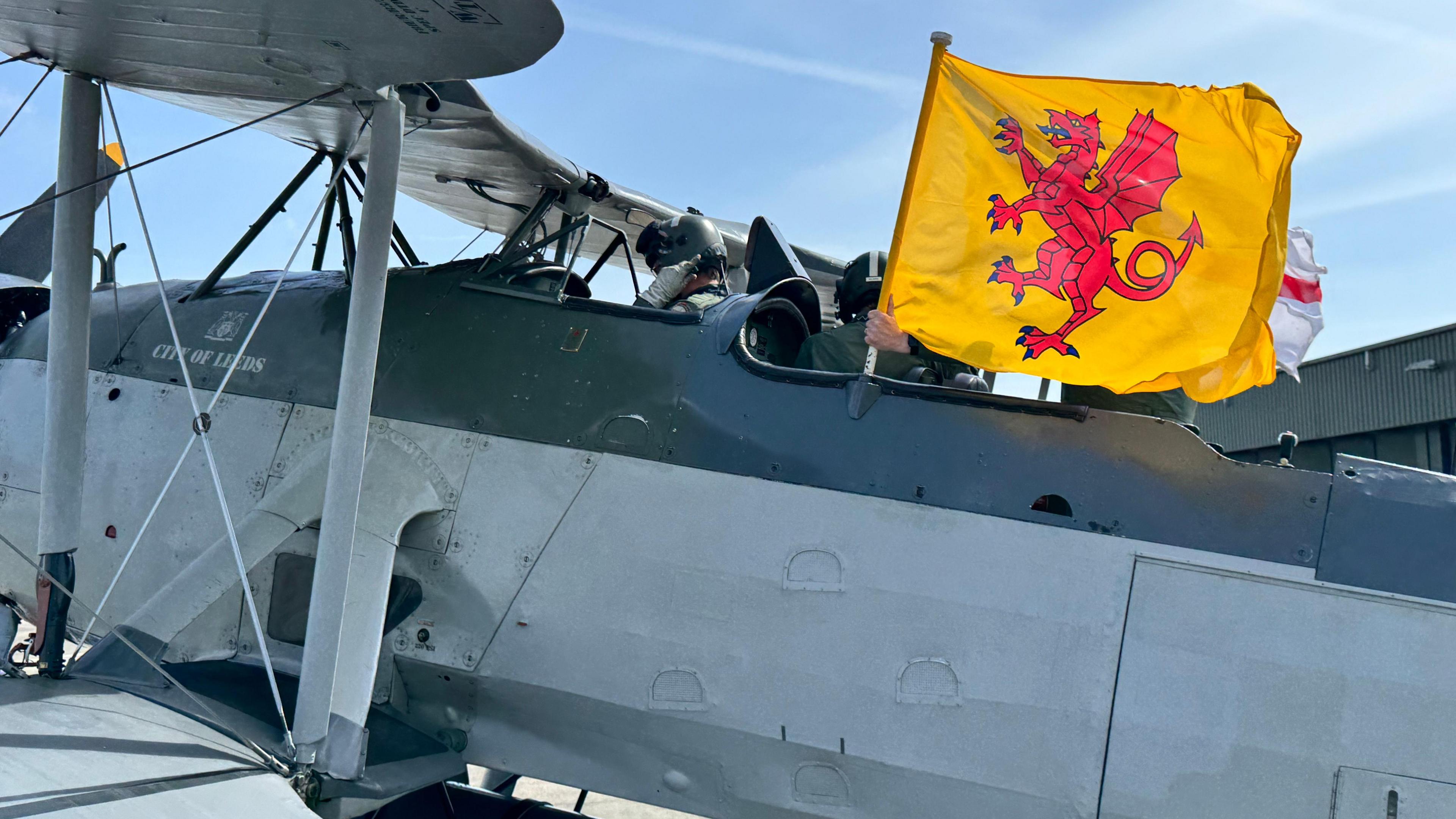 The outside of a small grey plane. The pilot is holding the yellow and red Somerset flag.