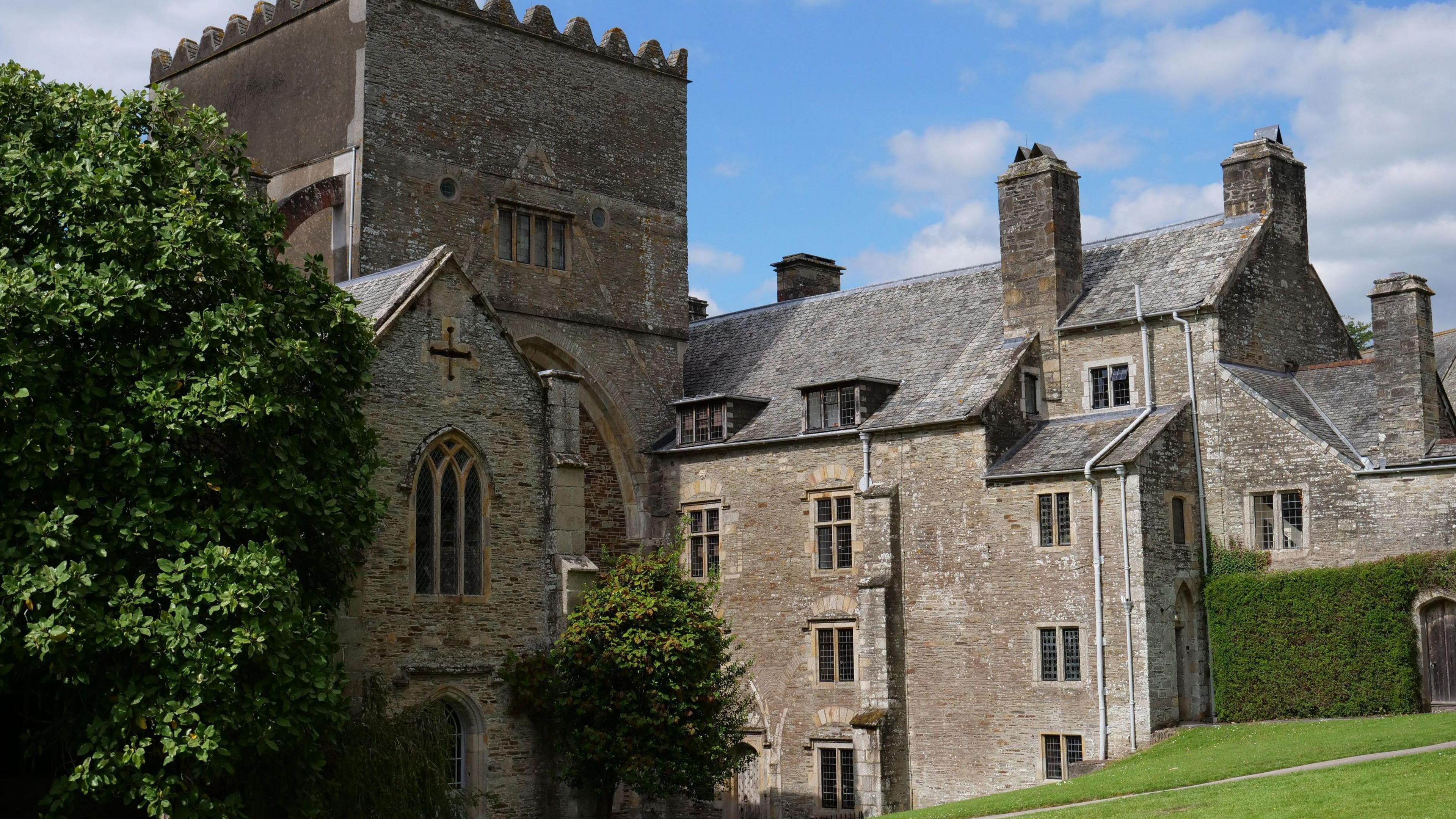 Buckland Abbey on a sunny day. It is a large grey bricked building with several levels and windows. Trees and a grass lawn are in front of the building.