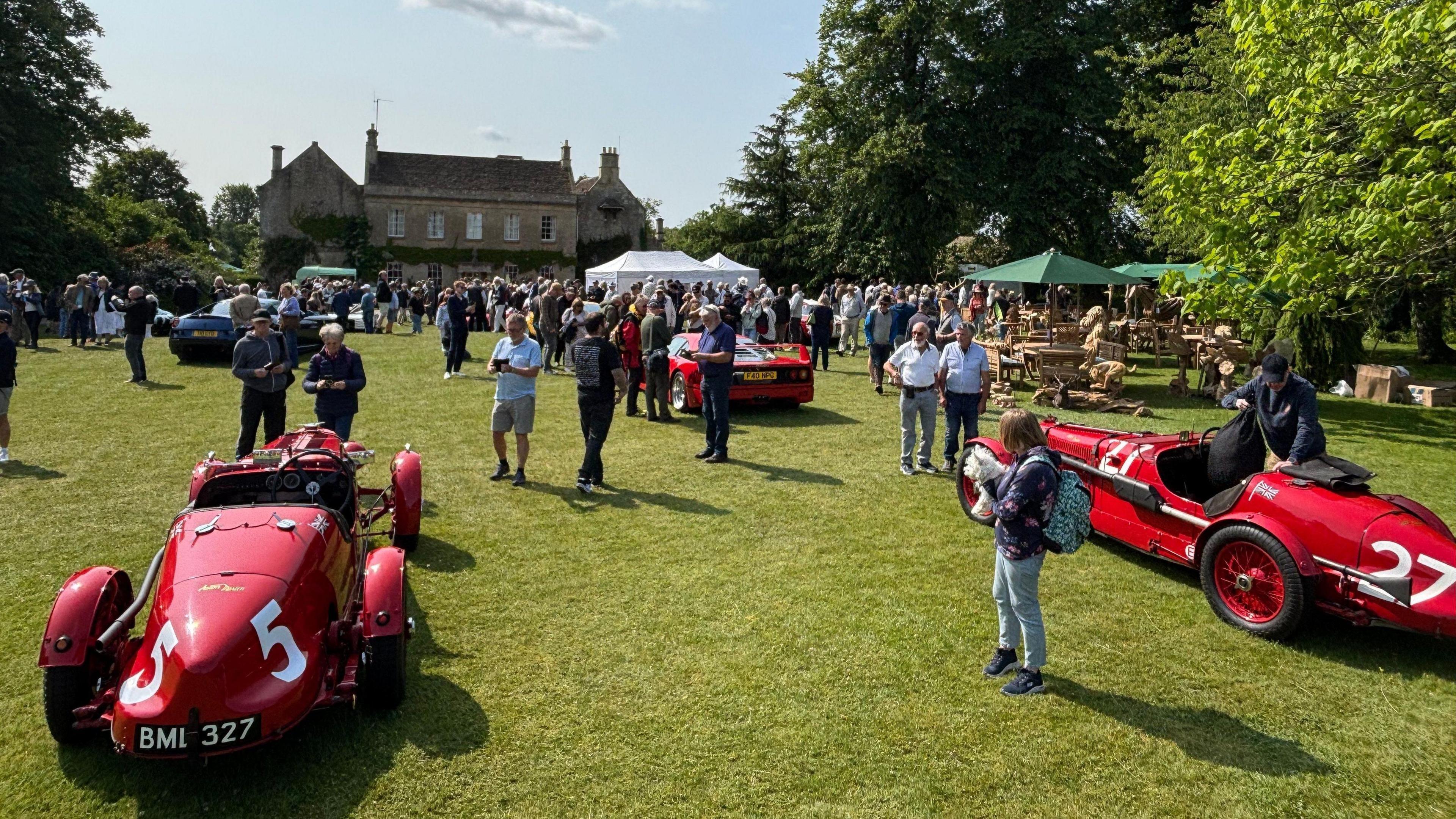An image showing the event Middlewick House. There are multiple red classic cars on display with a crowd of people on the field. There are also white and green stalls up