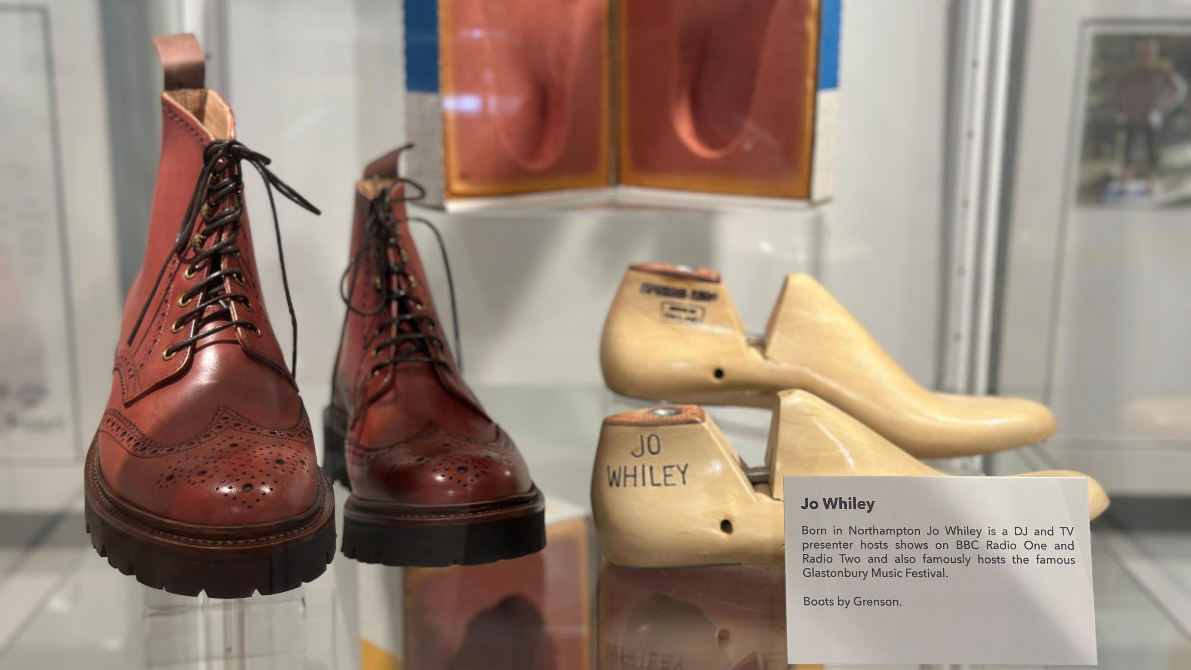 A closeup of brown brogue boots with laces and a pair of wooden lasts with Jo Whiley written on them. A white card with information about the shoes is next to them.