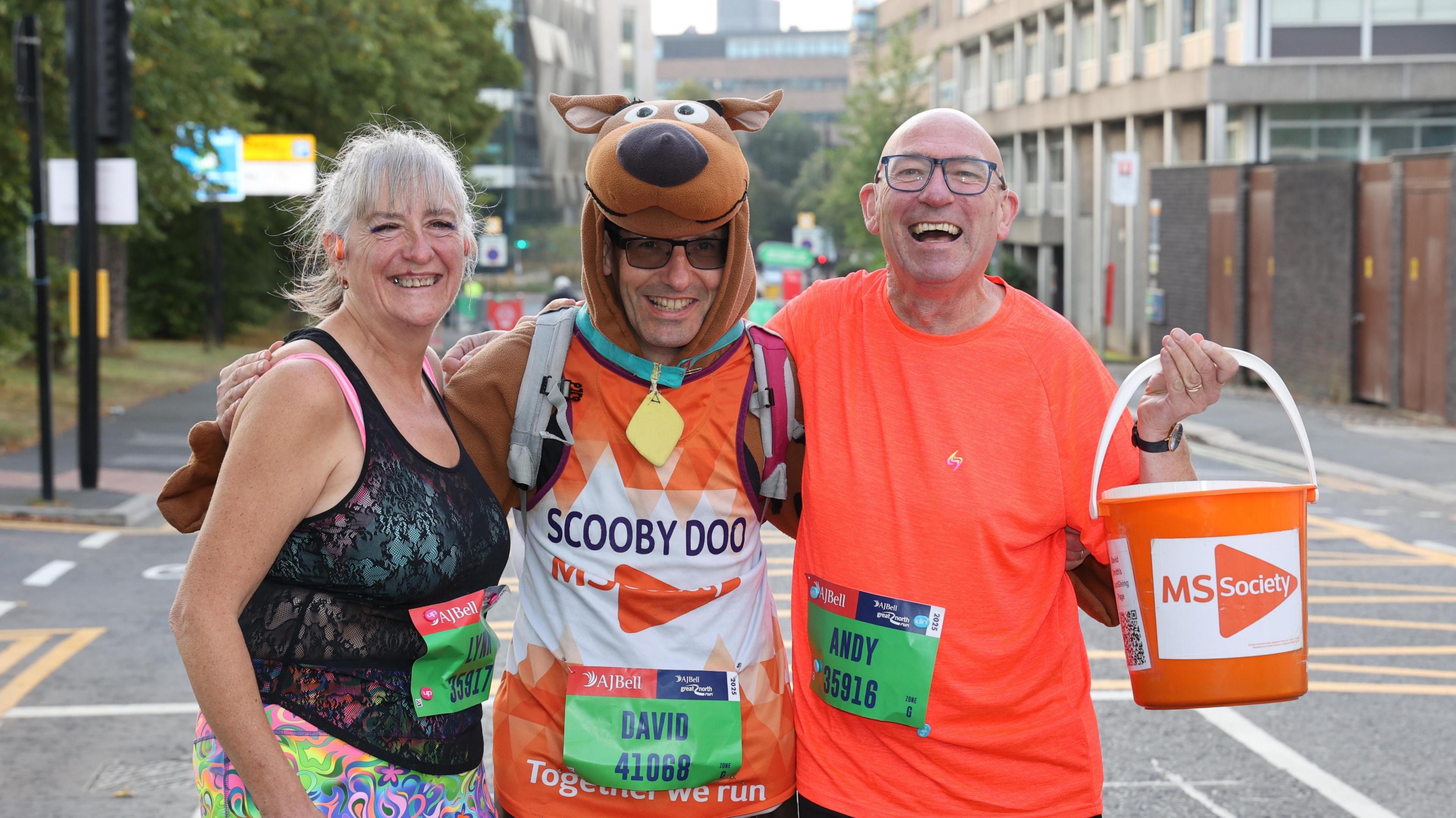 Three people in running gear smiling. The person in the middle is wearing a Scooby Doo costume.