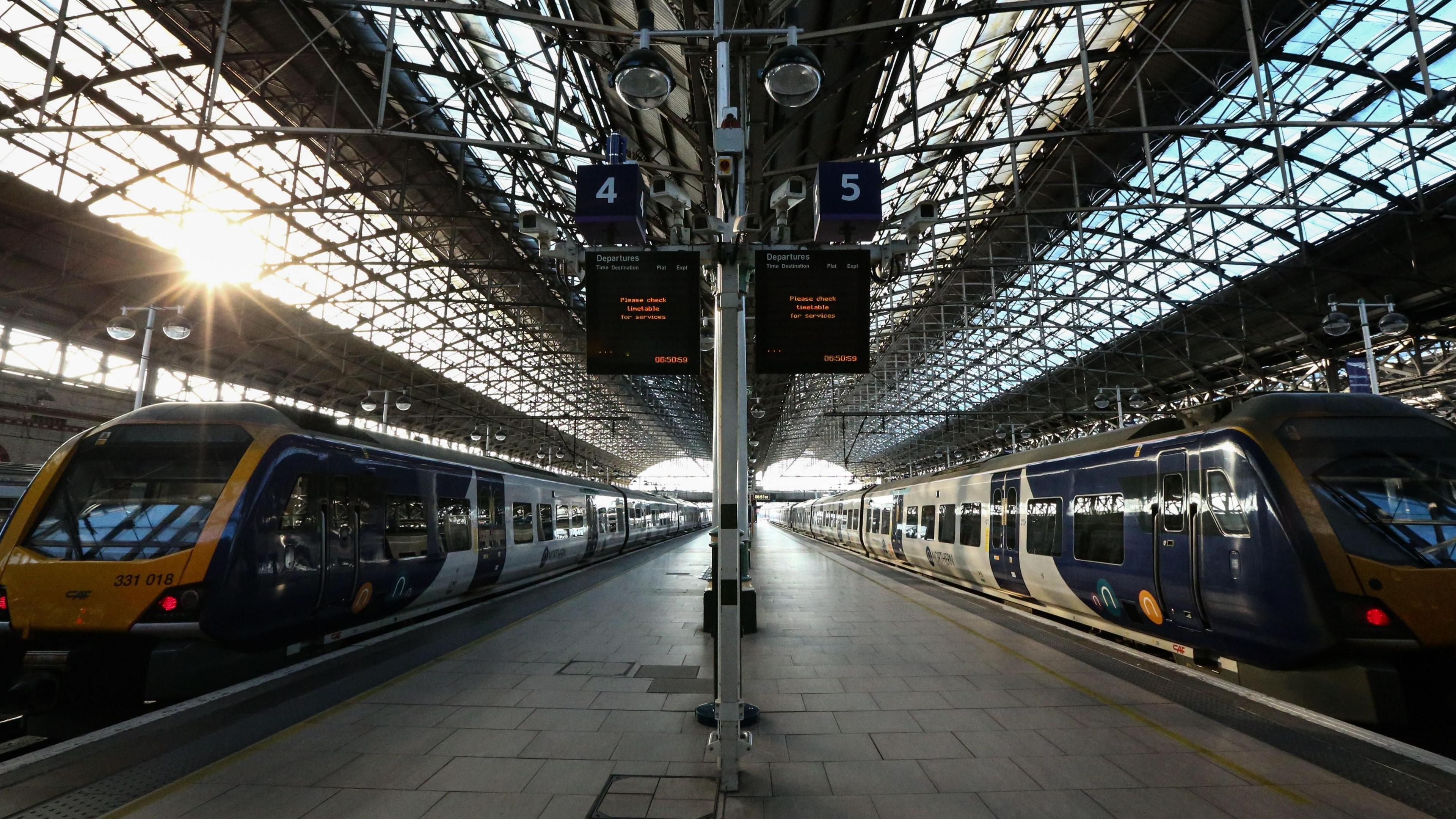 The interior of a large train station with two modern trains positioned on opposite platforms. The trains are sleek and mostly dark blue with yellow fronts, and they face toward the camera from the left and right sides of the image. The station has a high, arched glass roof supported by a metal framework, allowing natural light to stream in, creating bright reflections on the floor. In the centre of the image, there is a tall pole with two blue signs displaying platform numbers 4 and 5, and an electronic departure board showing train times in orange text. The platforms are wide and paved with light grey tiles, and the overall scene appears clean and spacious.