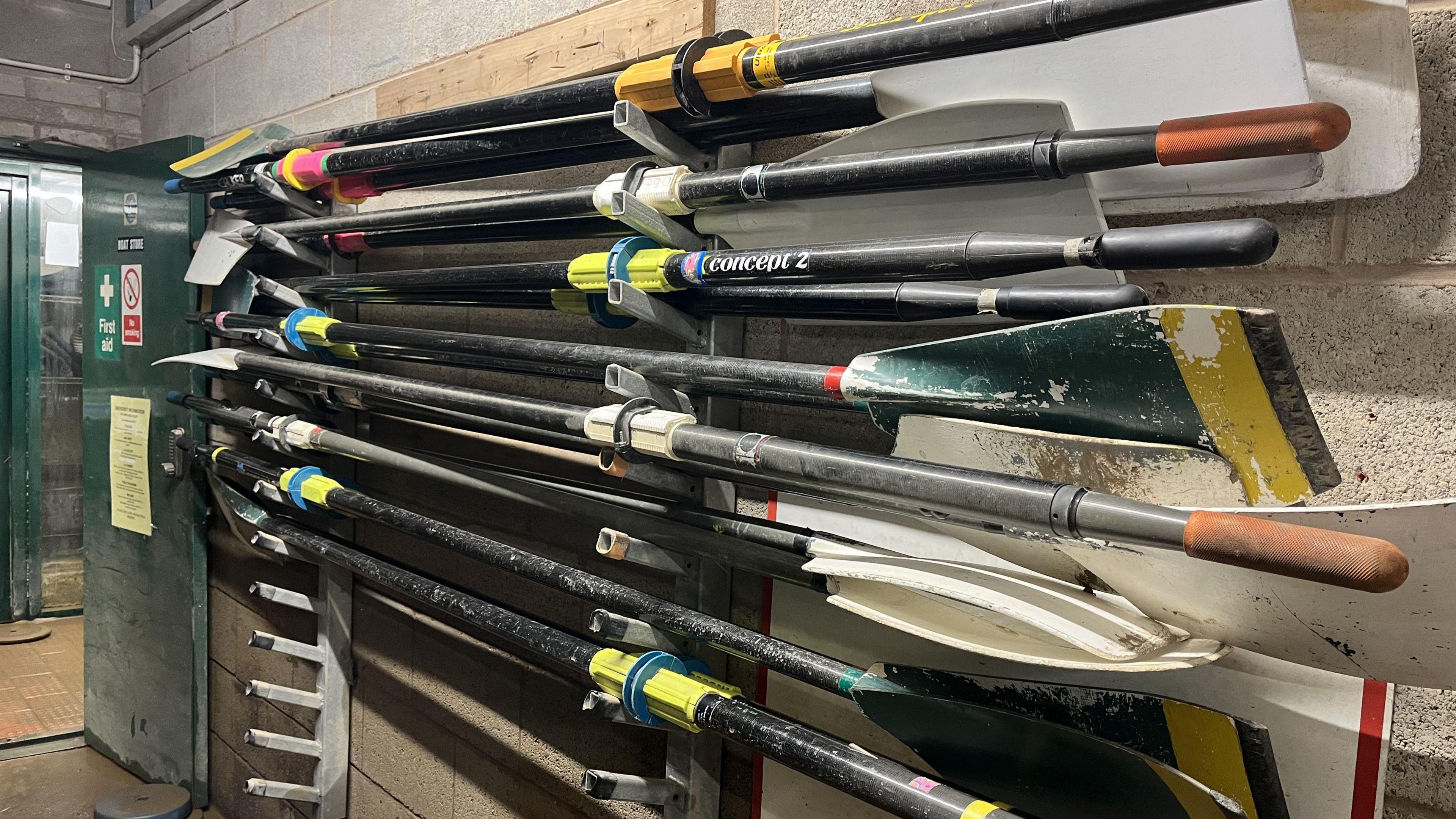 Several oars are stacked on small metal shelving on the wall of a clubhouse. They are covered in dirt and mud from flood water