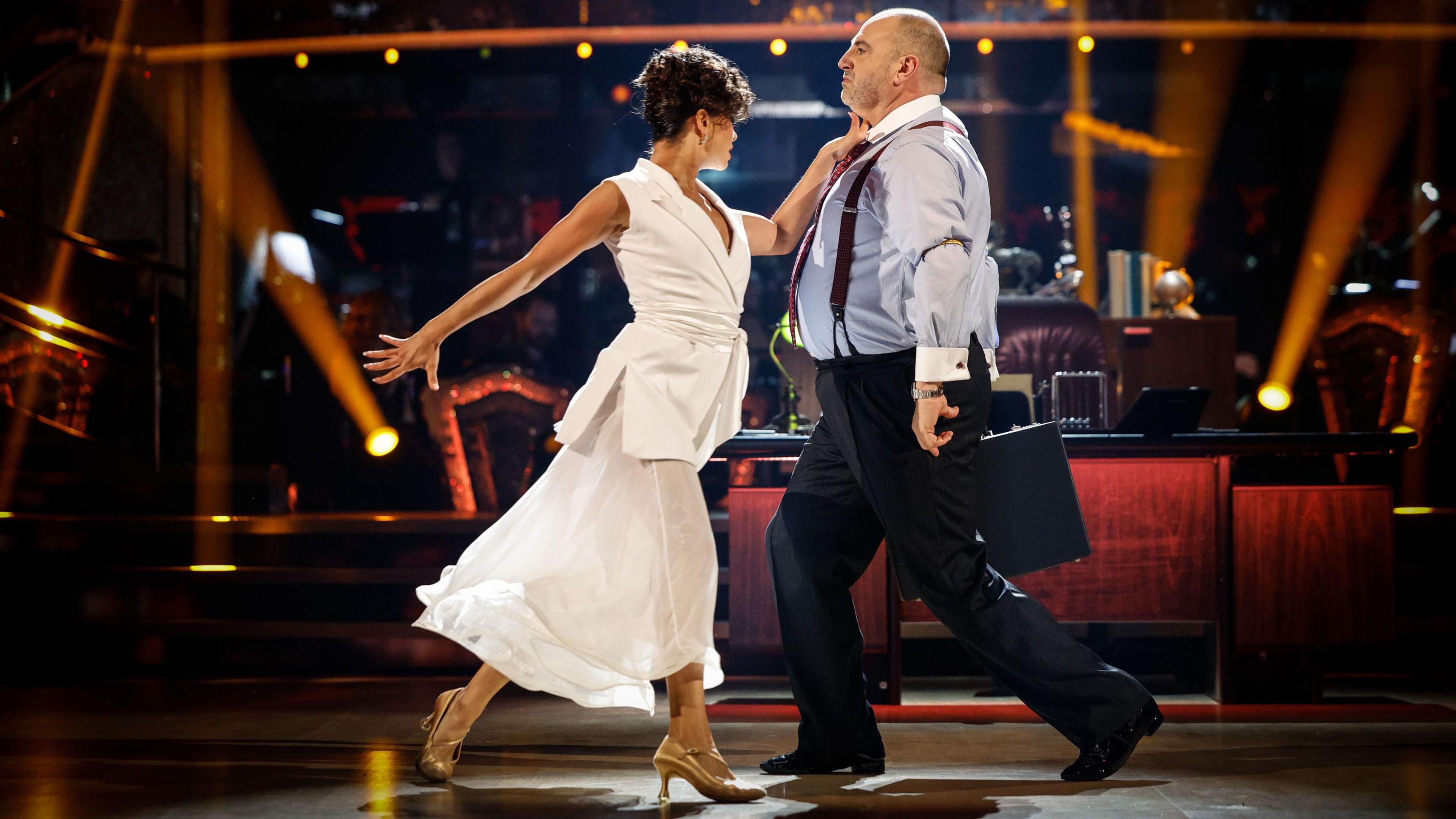 Wynne Evans, wearing a shirt and tie, and Katya Jones, wearing a white sleeveless blazer and skirt, dancing on the Strictly Come Dancing dancefloor.