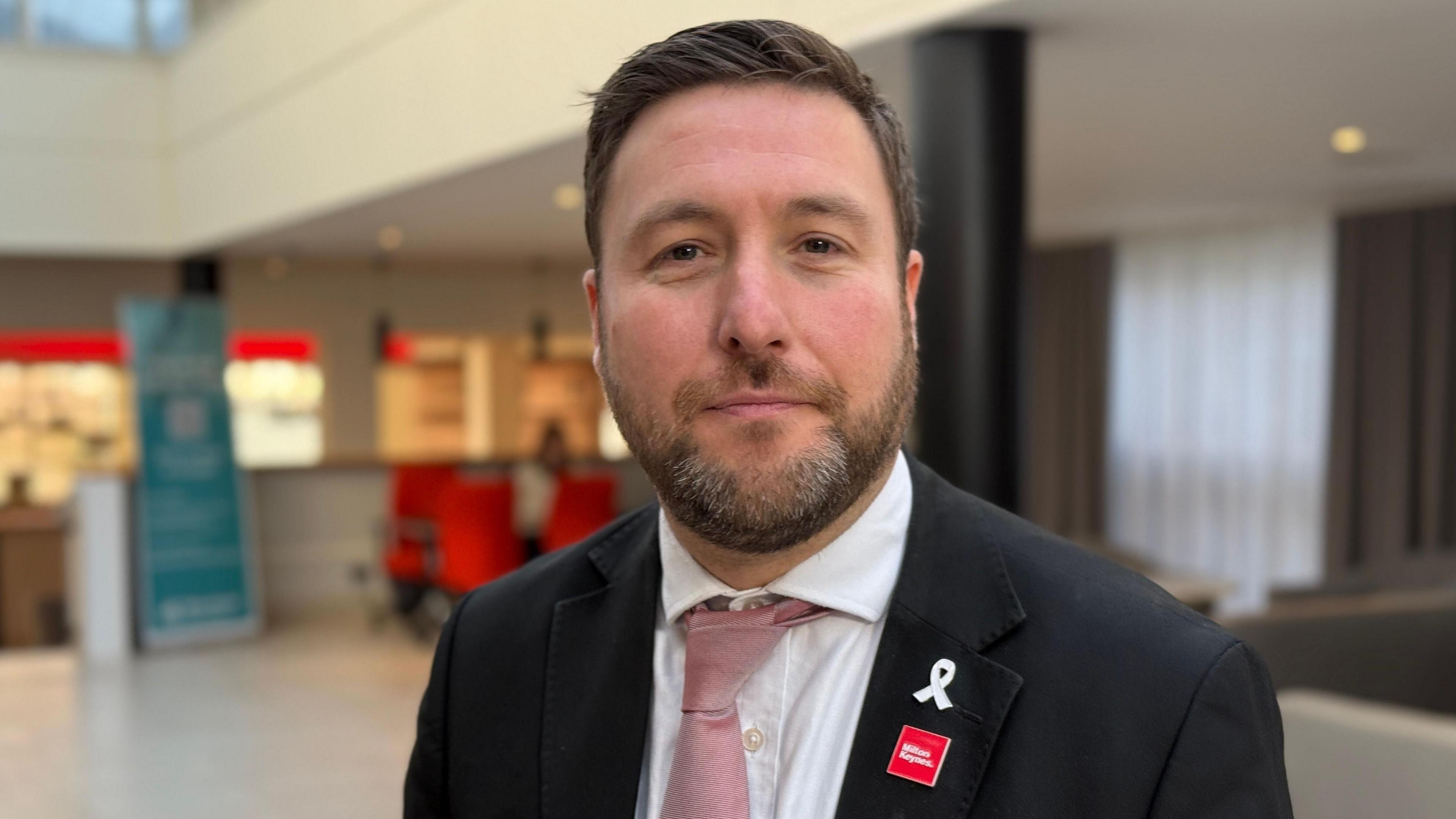 A picture of a man with short brown hair and a short goattee beard. He is wearing a black suit jacket and has a pink tie and white shirt on.