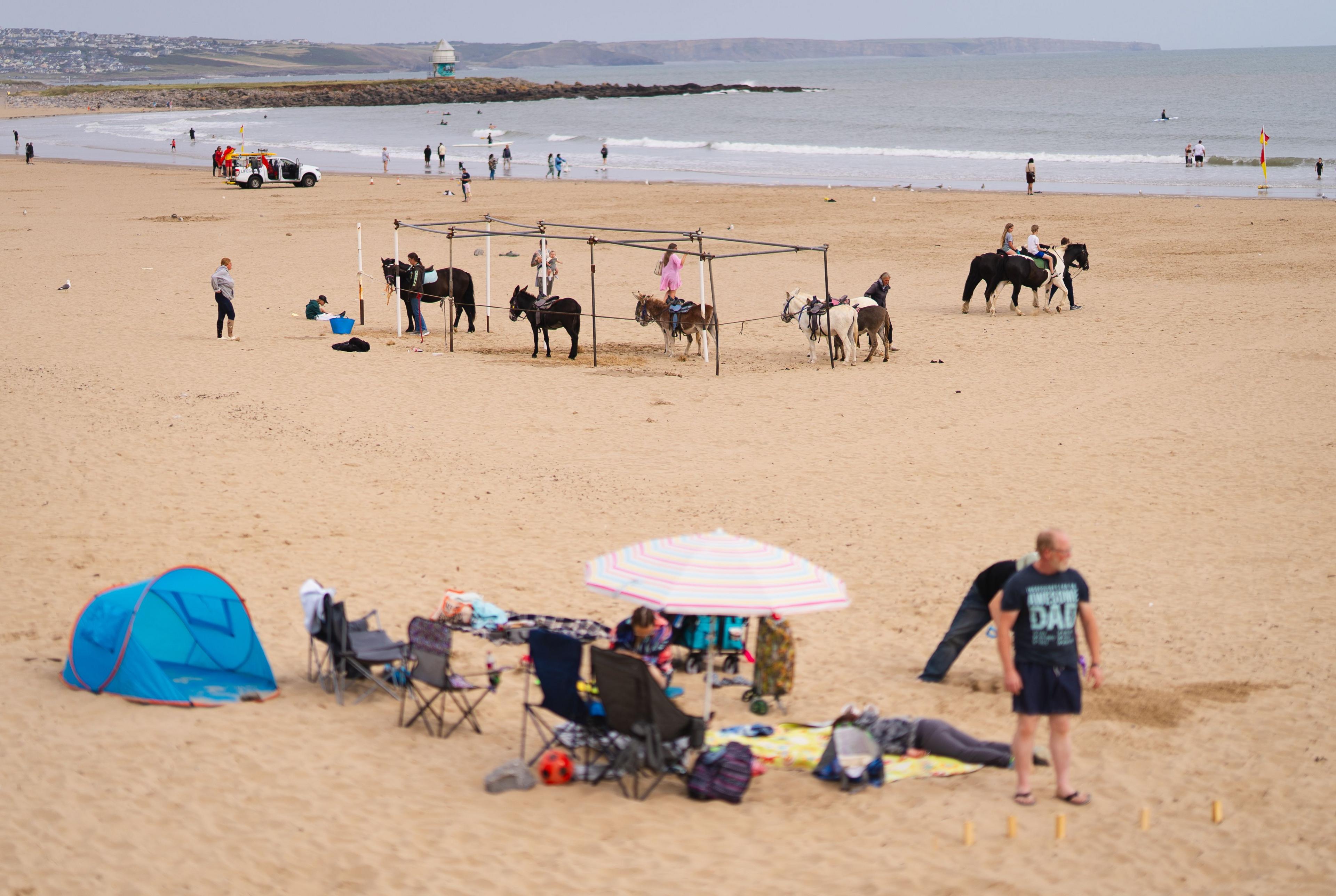 Children on donkeys on a sandy beach with a breakwater in the distance. A family with beach tent, deck chairs and parasol are relaxing on the beach in the foreground.