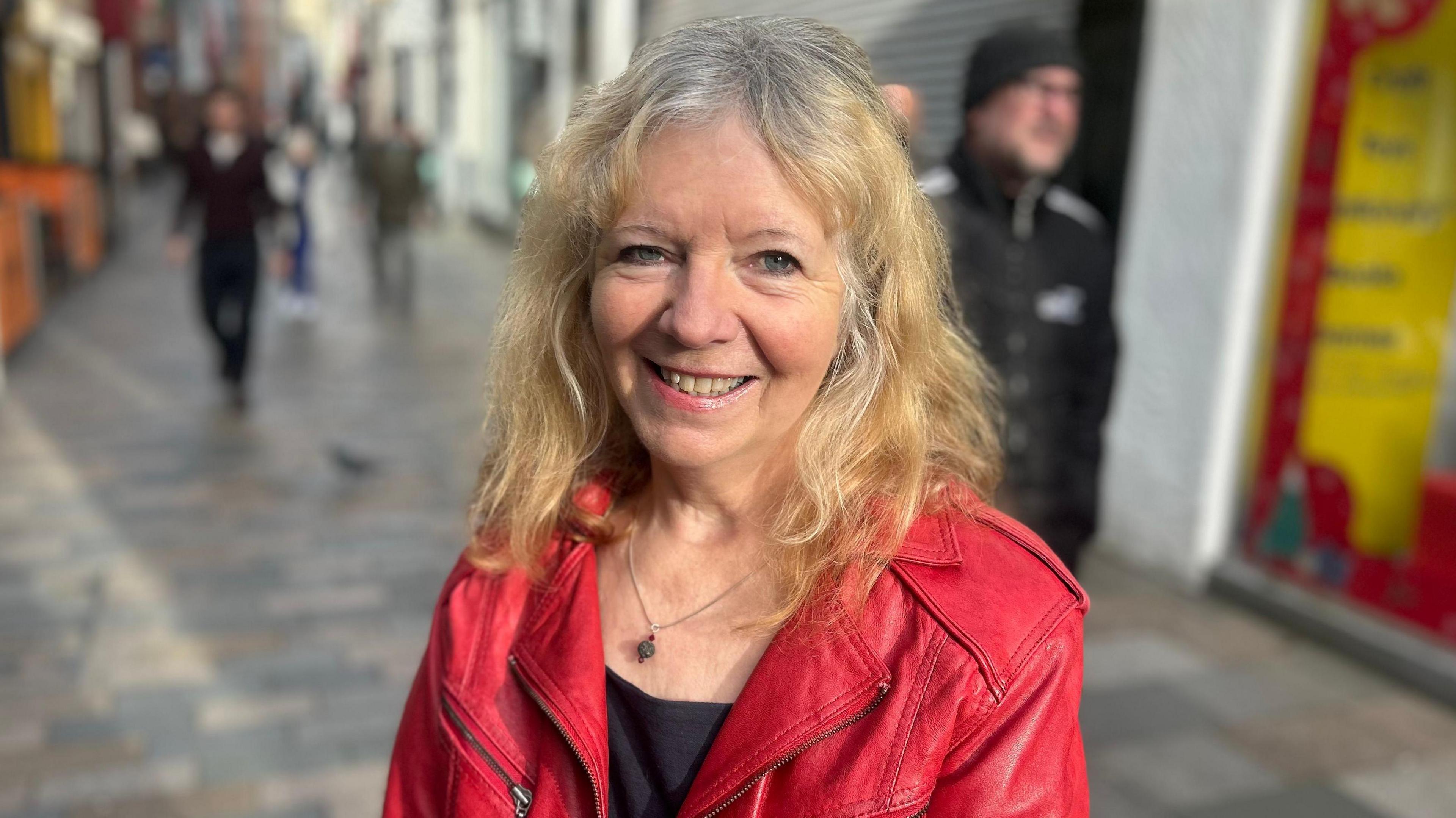 Lucia Shepherd standing in Strand Street, which is lined with shops, smiling at the camera. She has blonde curly hair and is wearing a red leather jacket.