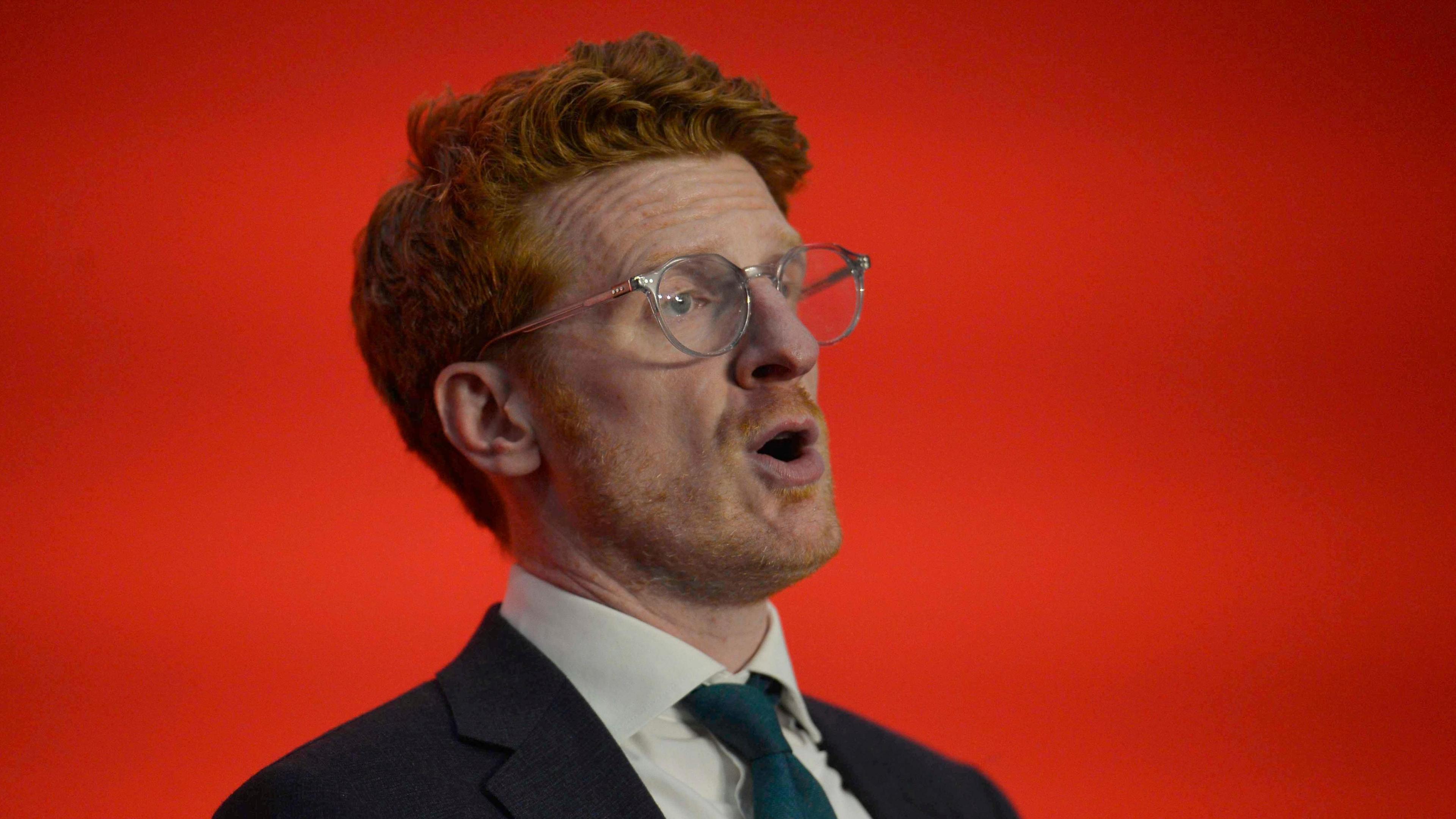 Matthew is mid speech in front of a bold red background. 
He is wearing silver small rimmed glasses with a pink arm. 
He has ginger curly short hair.
He's wearing a teal blue tie, white shirt and navy suit blazer. 
