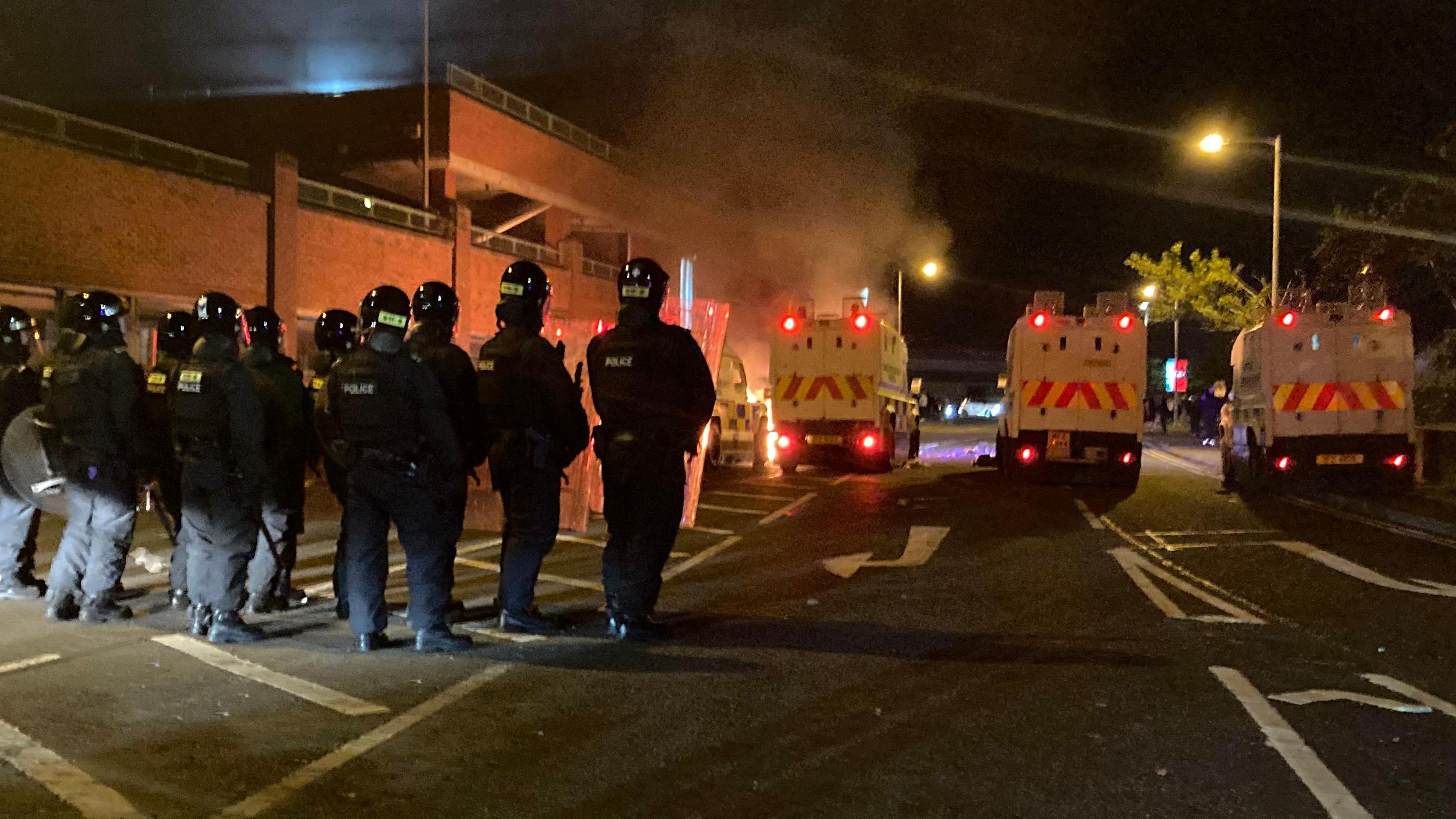 Police in riot gear with their backs to the camera. There are police land rovers in front of them in a row. In front of the land rovers there is fire and smoke.