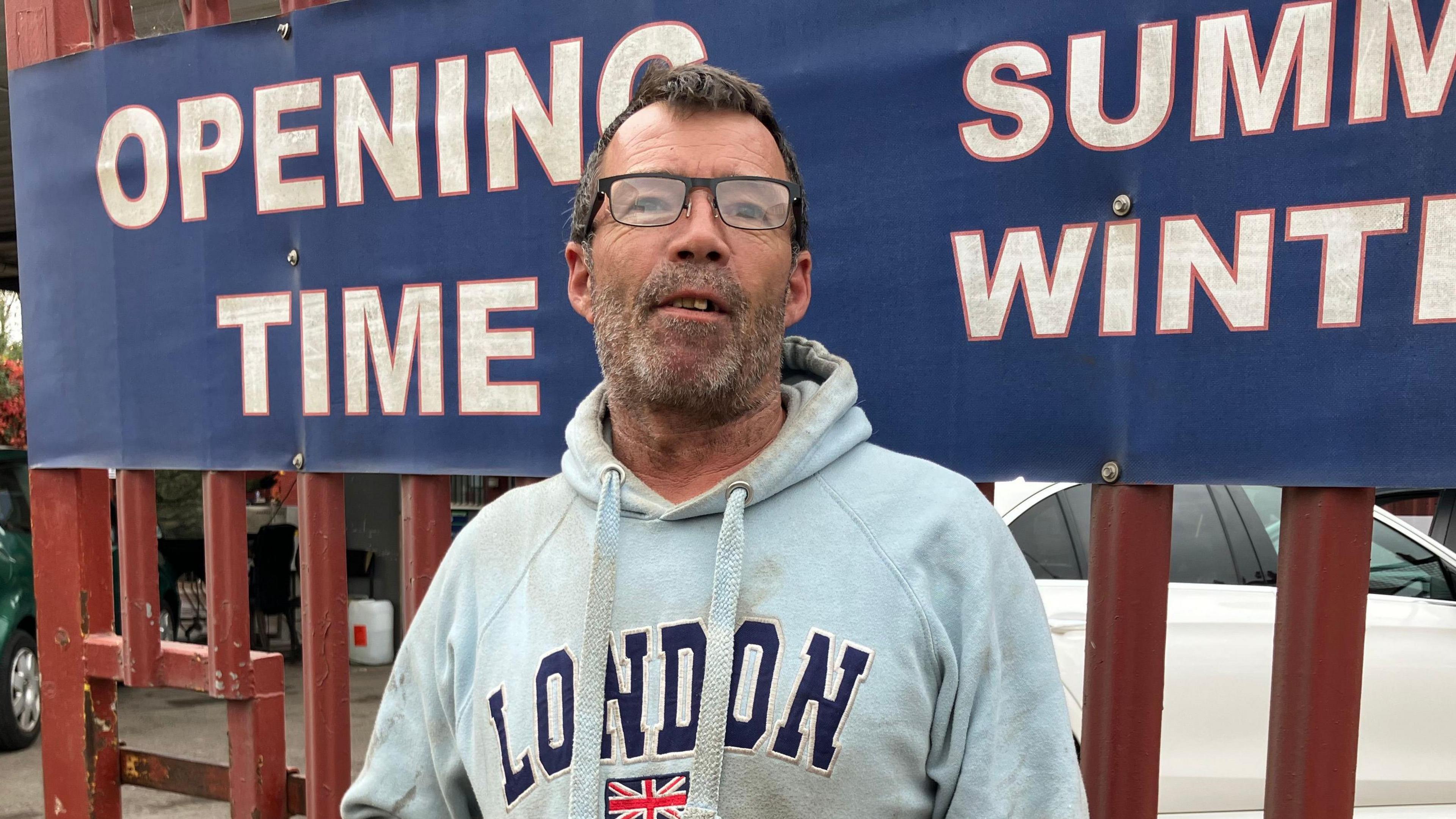 A man wearing a hoodie and looking at the camera. He is wearing black frame glasses. His hoodie is pale blue and says London on it