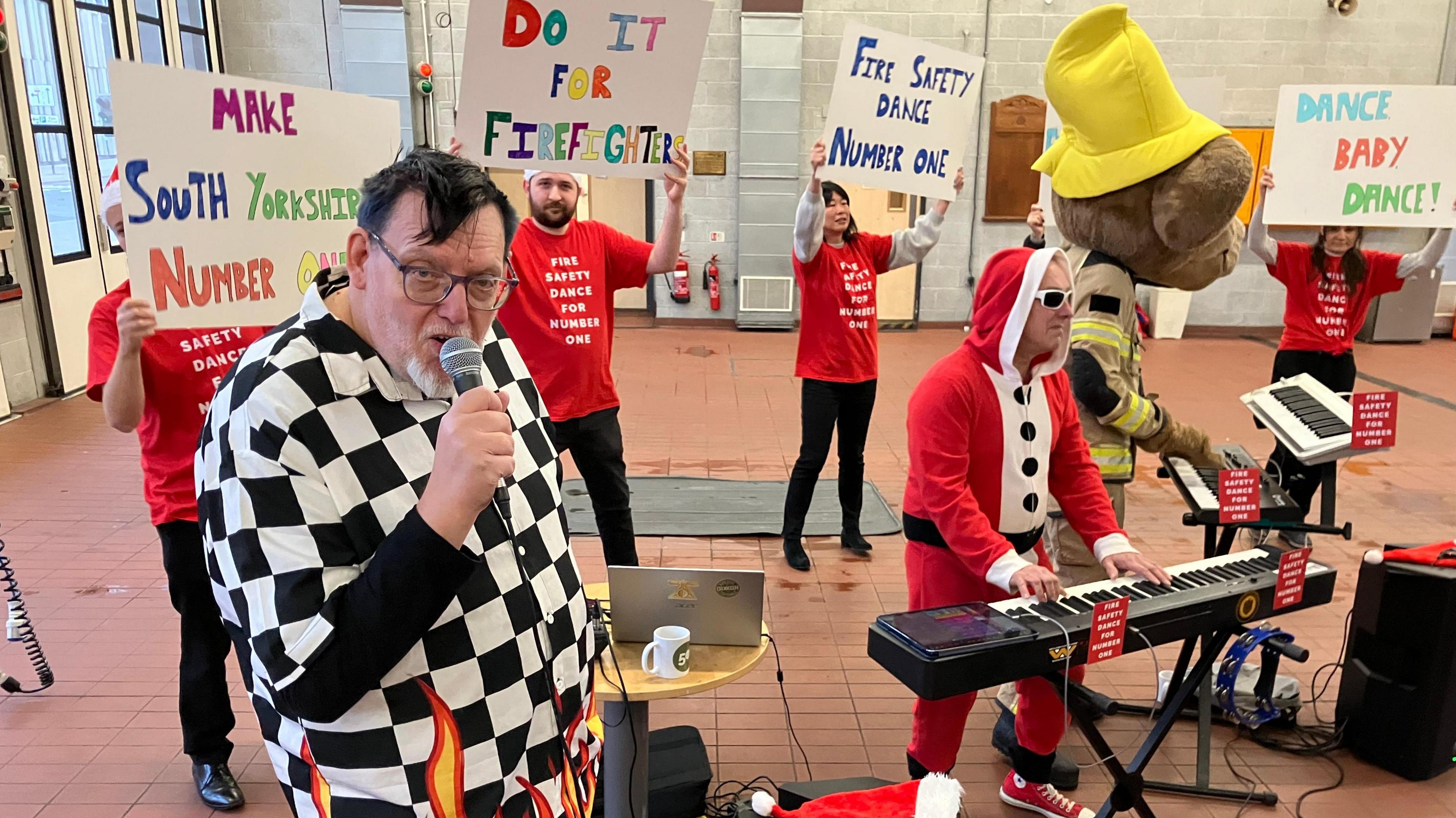 A man in a black and white checked shirt sings into a microphone, while a man in a Santa onesie plays the keyboard and others in red t shirts hold up signs.