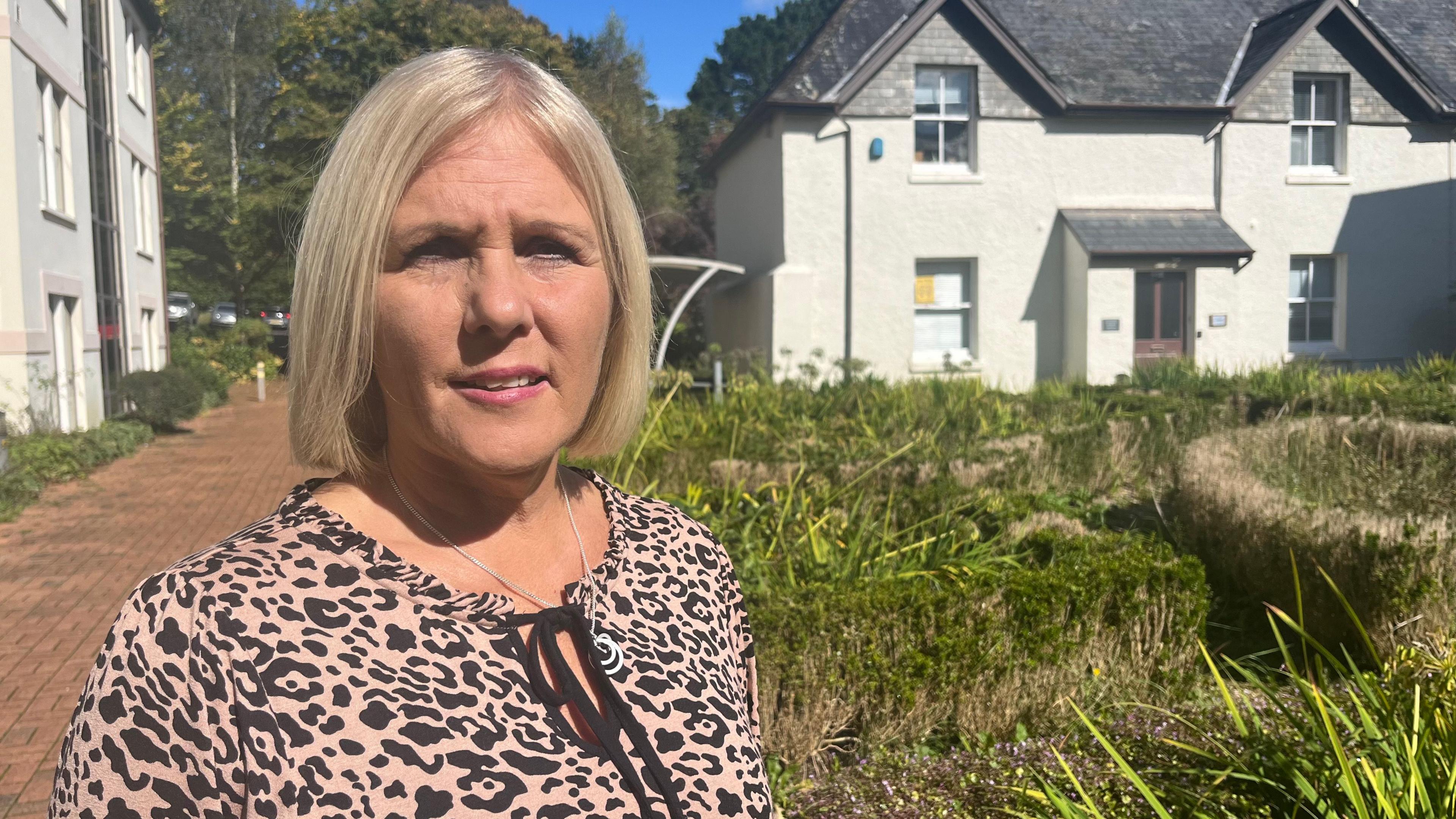 Councillor Nicky Hopwood, wearing an animal print top, standing in a grassy area with offices in the background. She has chin length blonde hari and is looking at the camer and wearing a silver pendant on a chain and pink lipstick