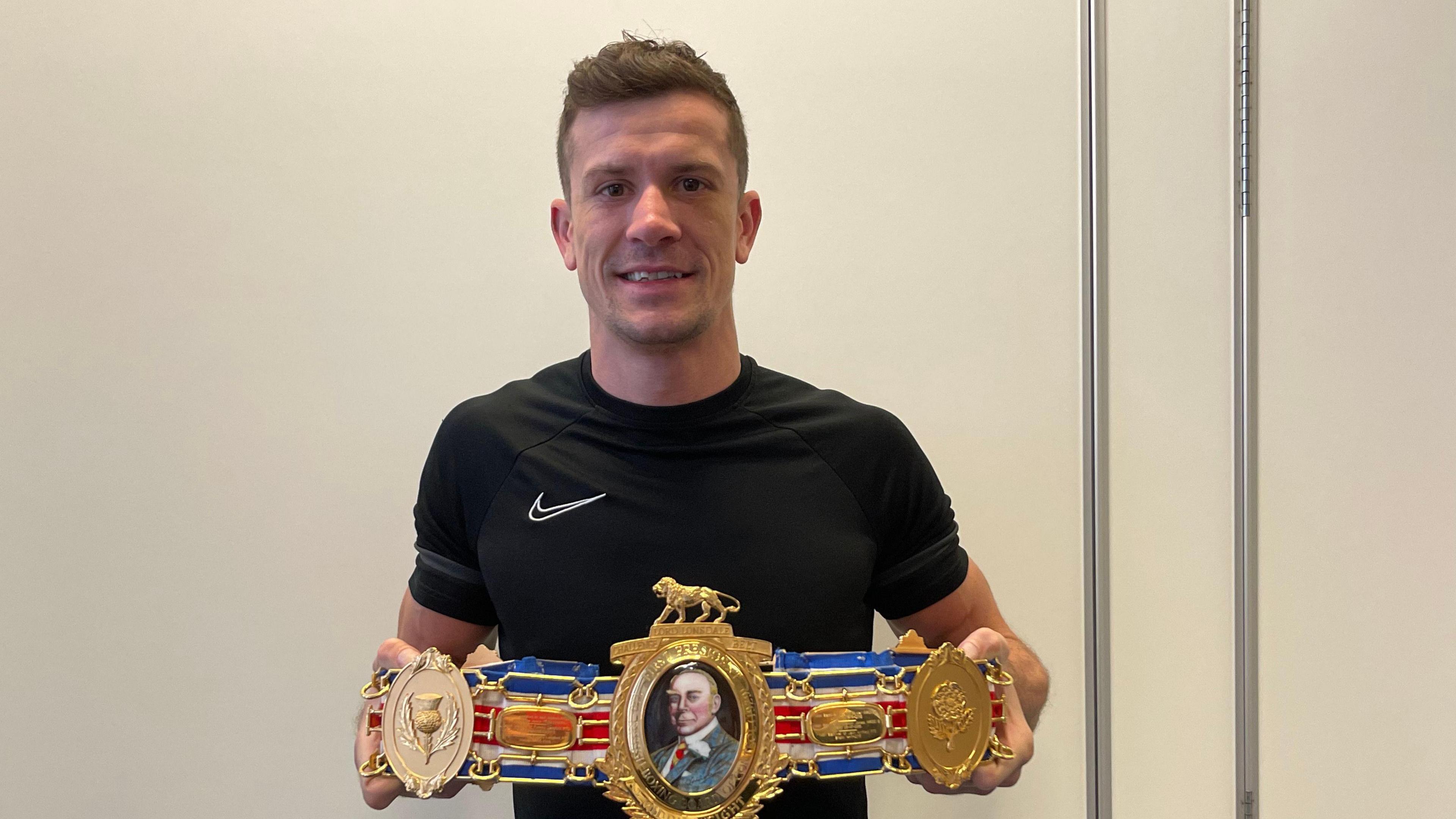 Nathan Heaney is stood wearing a black top, holding an ornate boxing belt. He is stood against a white background, which has folding partitions.