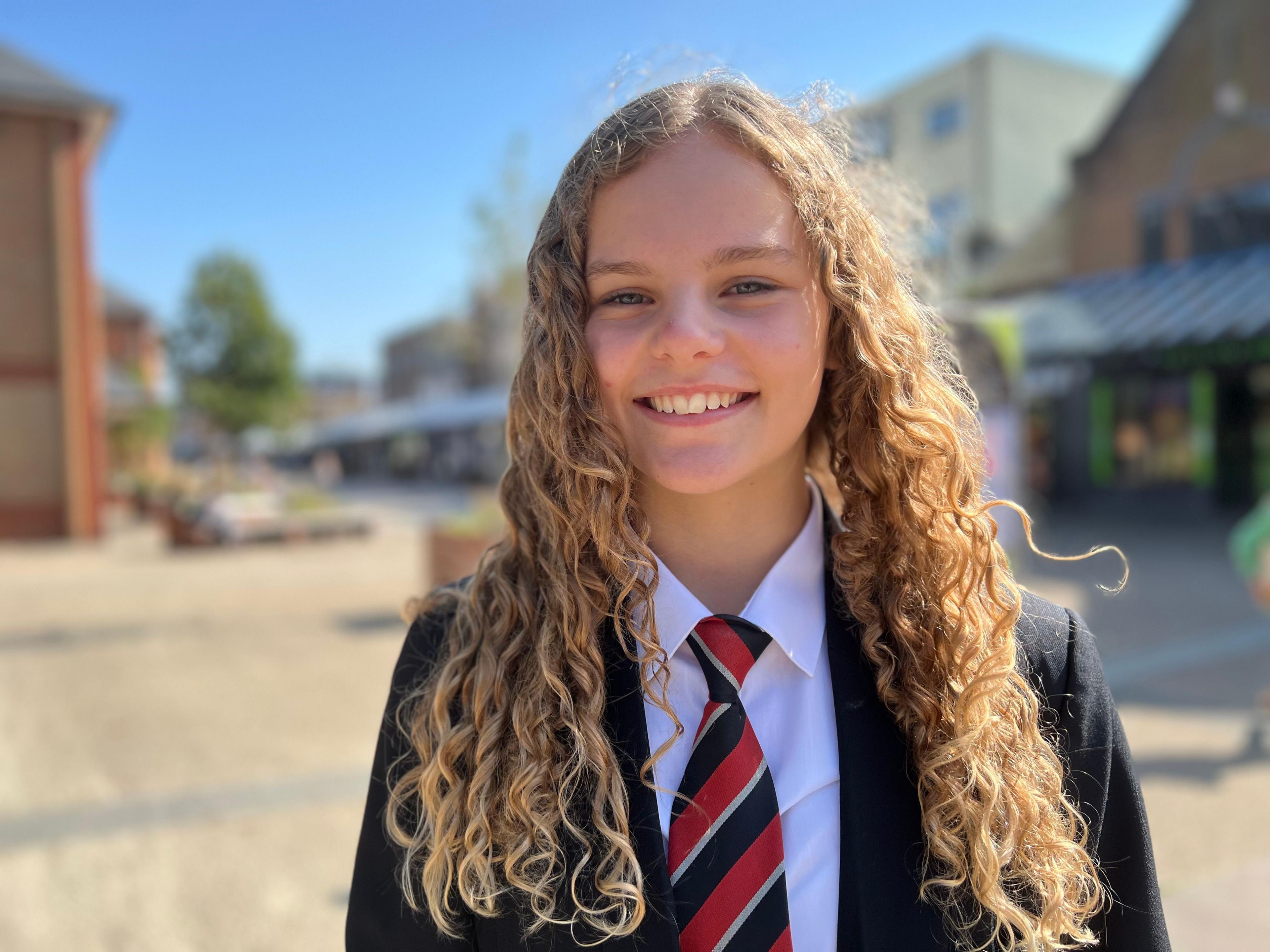 A head and shoulders shot of a girl in school uniform. She is wearing a red, black and white striped tie, a white shirt and black blazer. She has long, curly, blonde hair