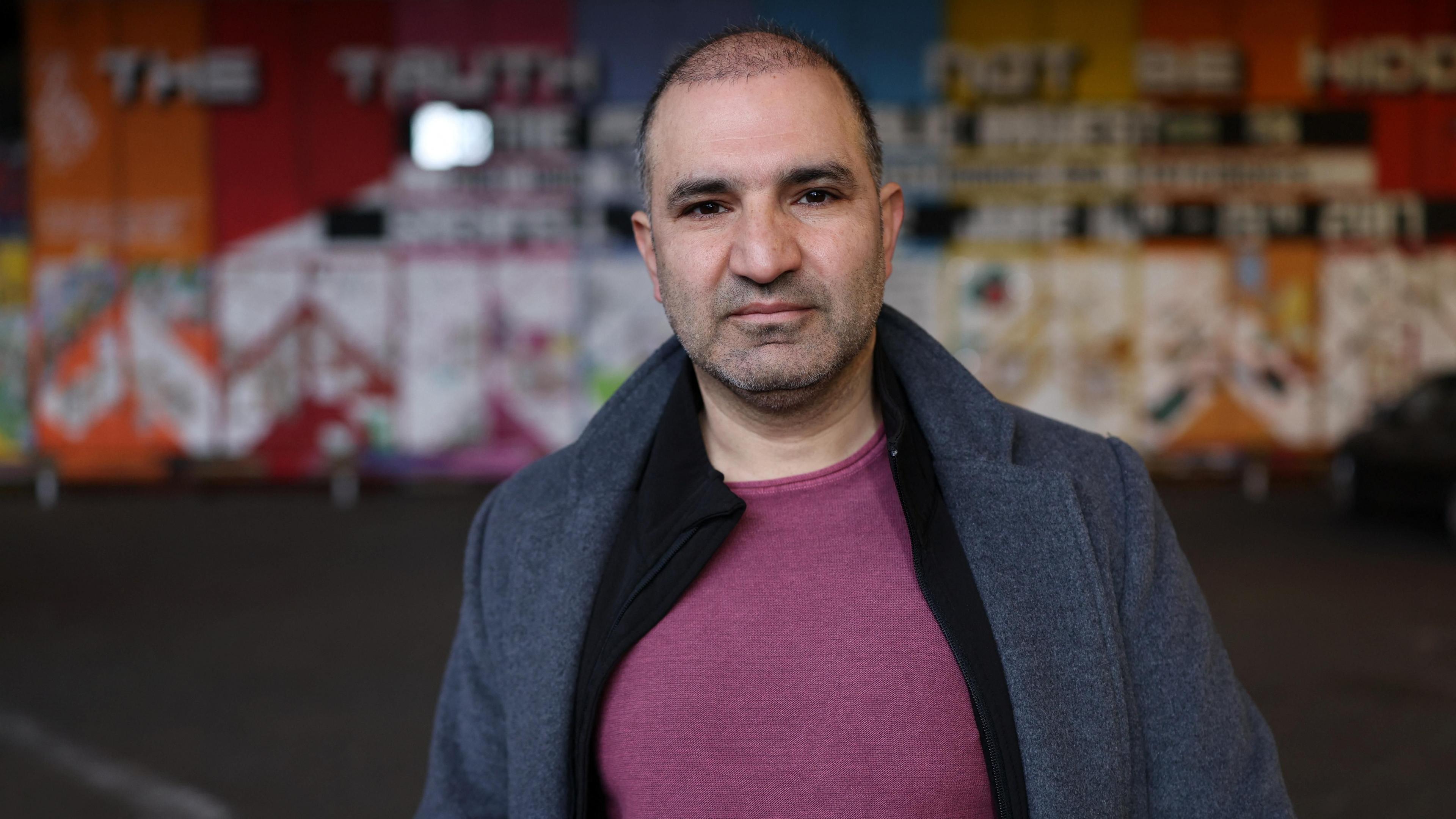 Hamid Ali Jafari, a man with black hair wearing a grey jacket and red top, stands in front of a colourful wall.