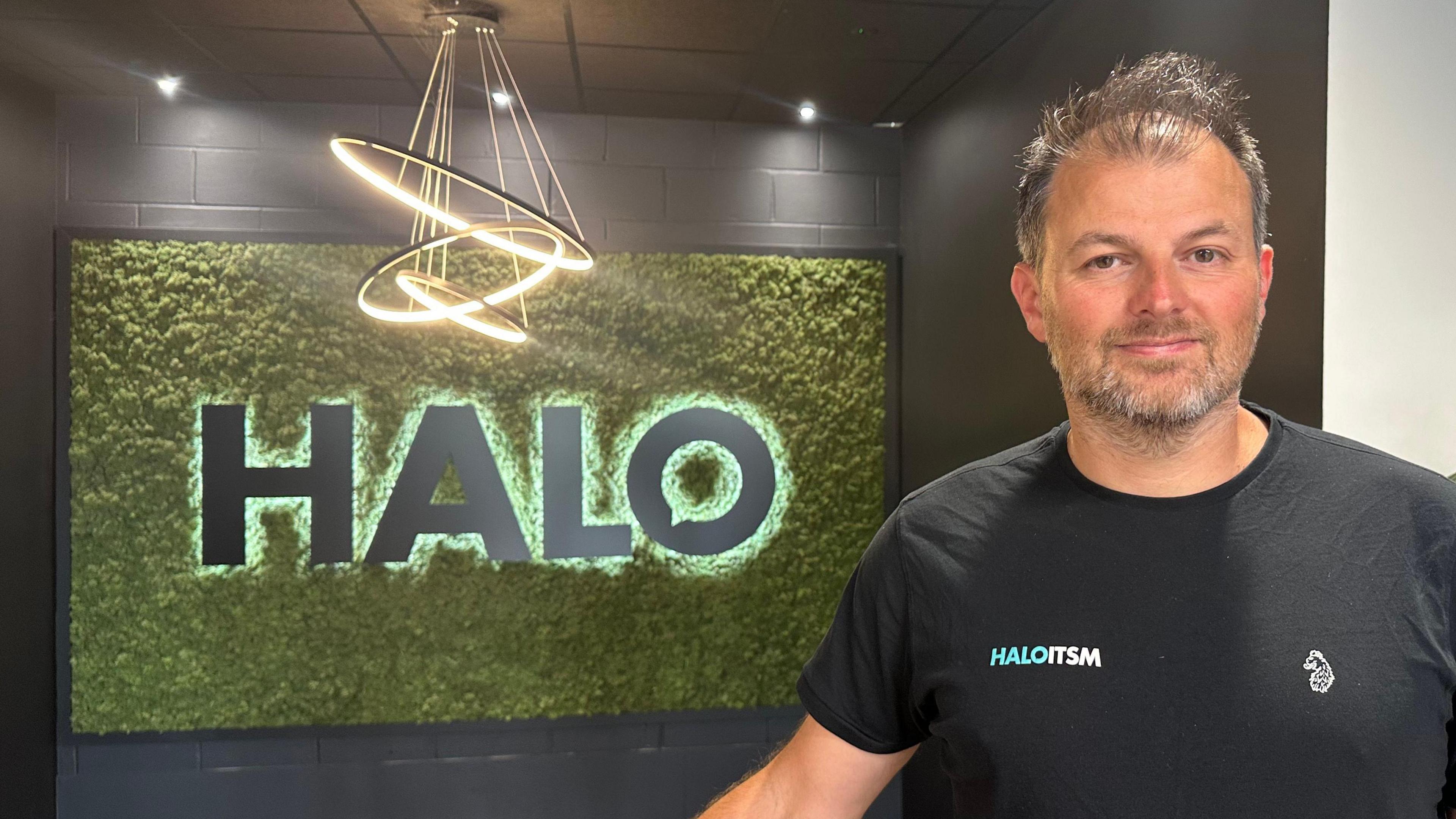 Paul Hamilton stands and smiles at the camera in front of a large sign that reads his company's name, Halo. He has grey/dark hair and wears a black T-shirt with his company's ITSM logo on it. 